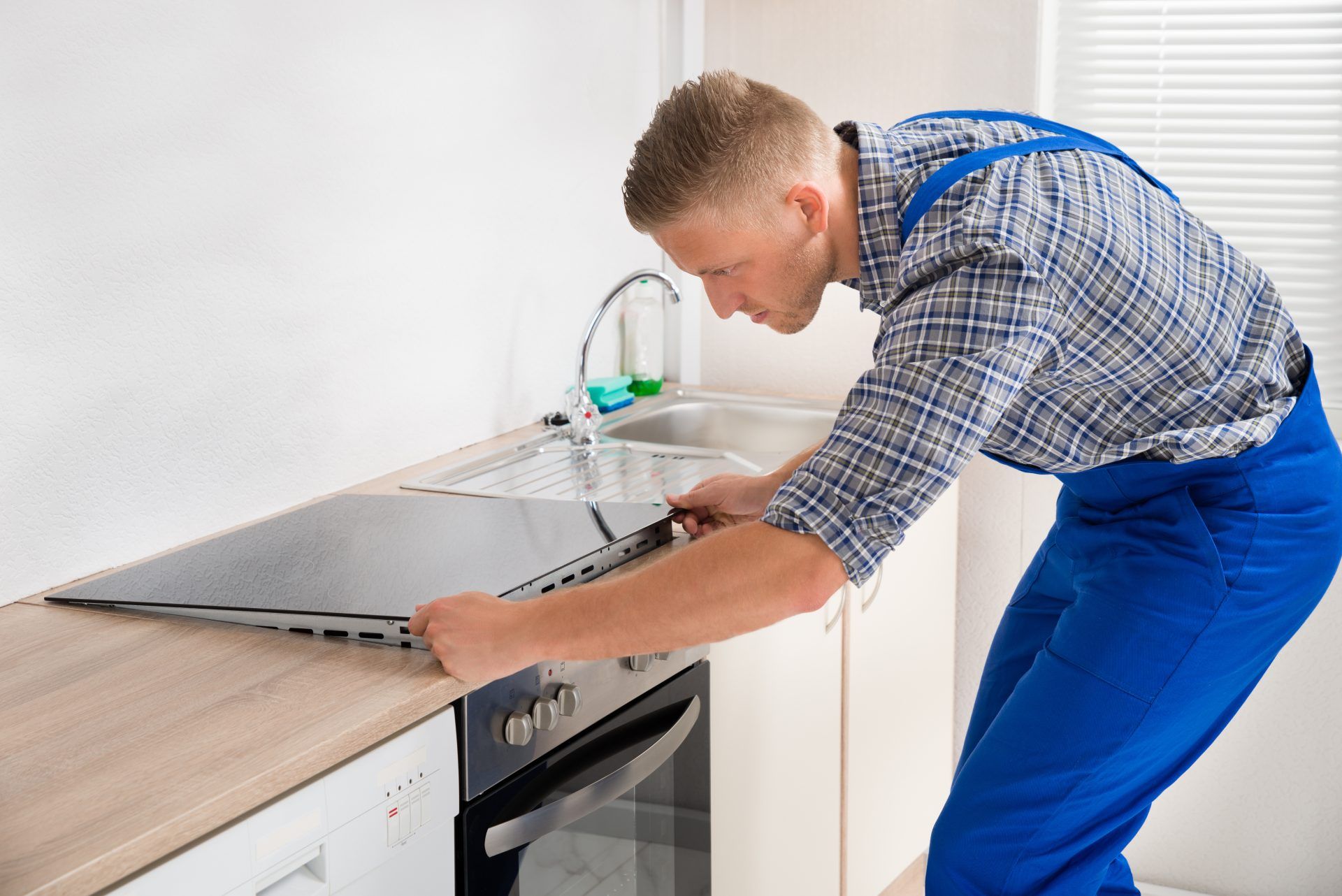 Man in blue overalls installing a black stovetop in a kitchen.