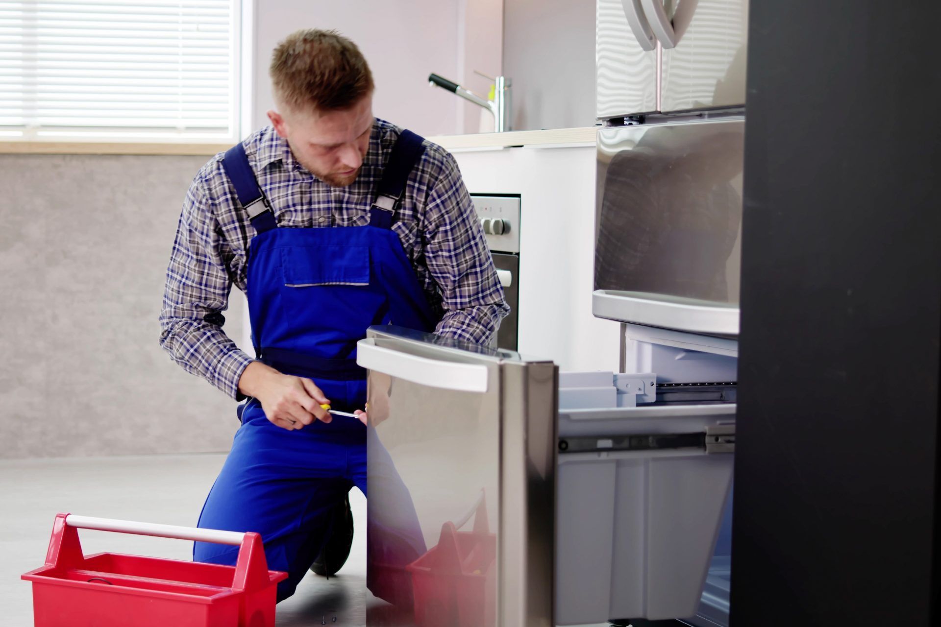 Person kneeling, fixing refrigerator with a screwdriver, red toolbox, kitchen setting.