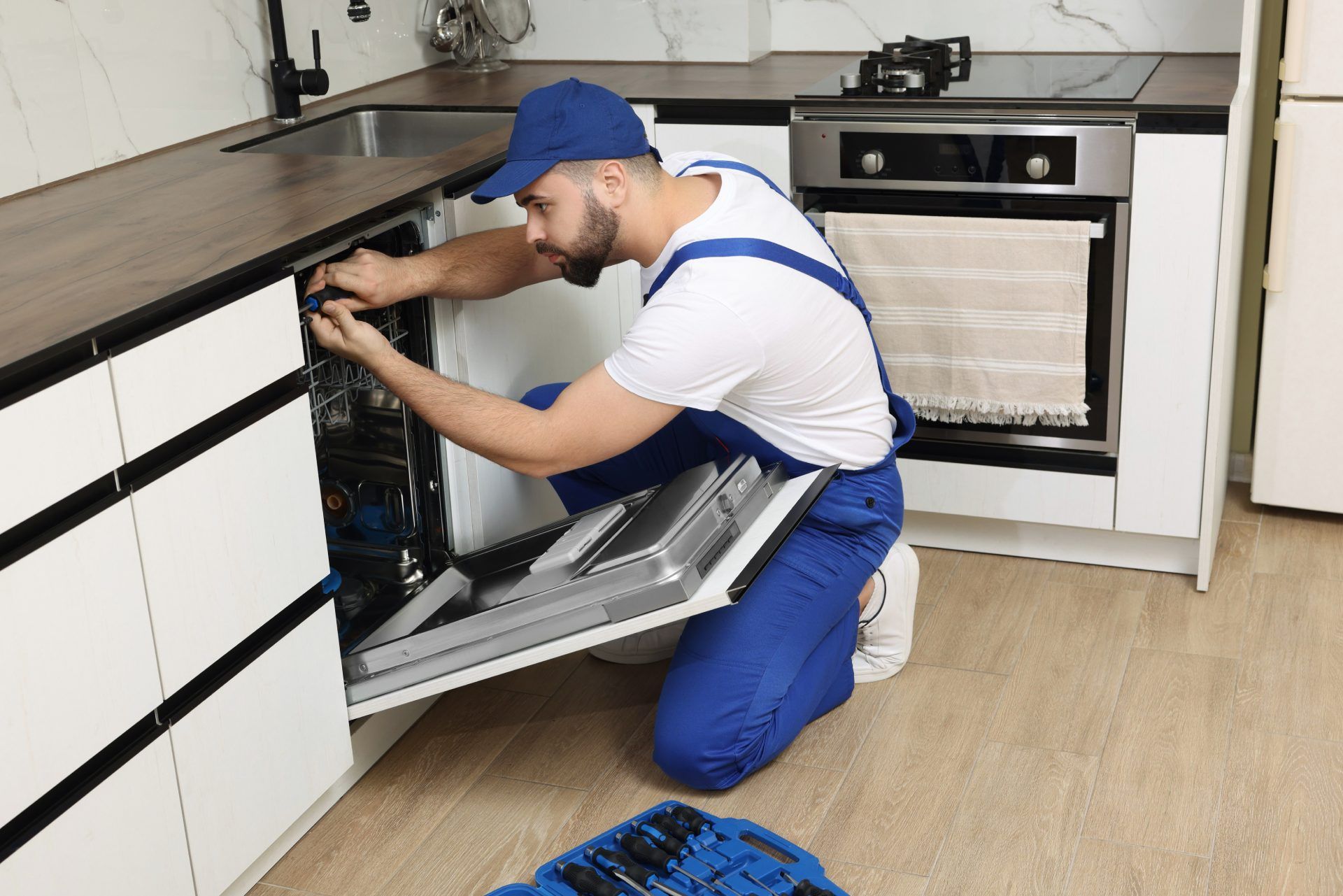 Man in blue overalls and cap repairs a dishwasher in a kitchen.