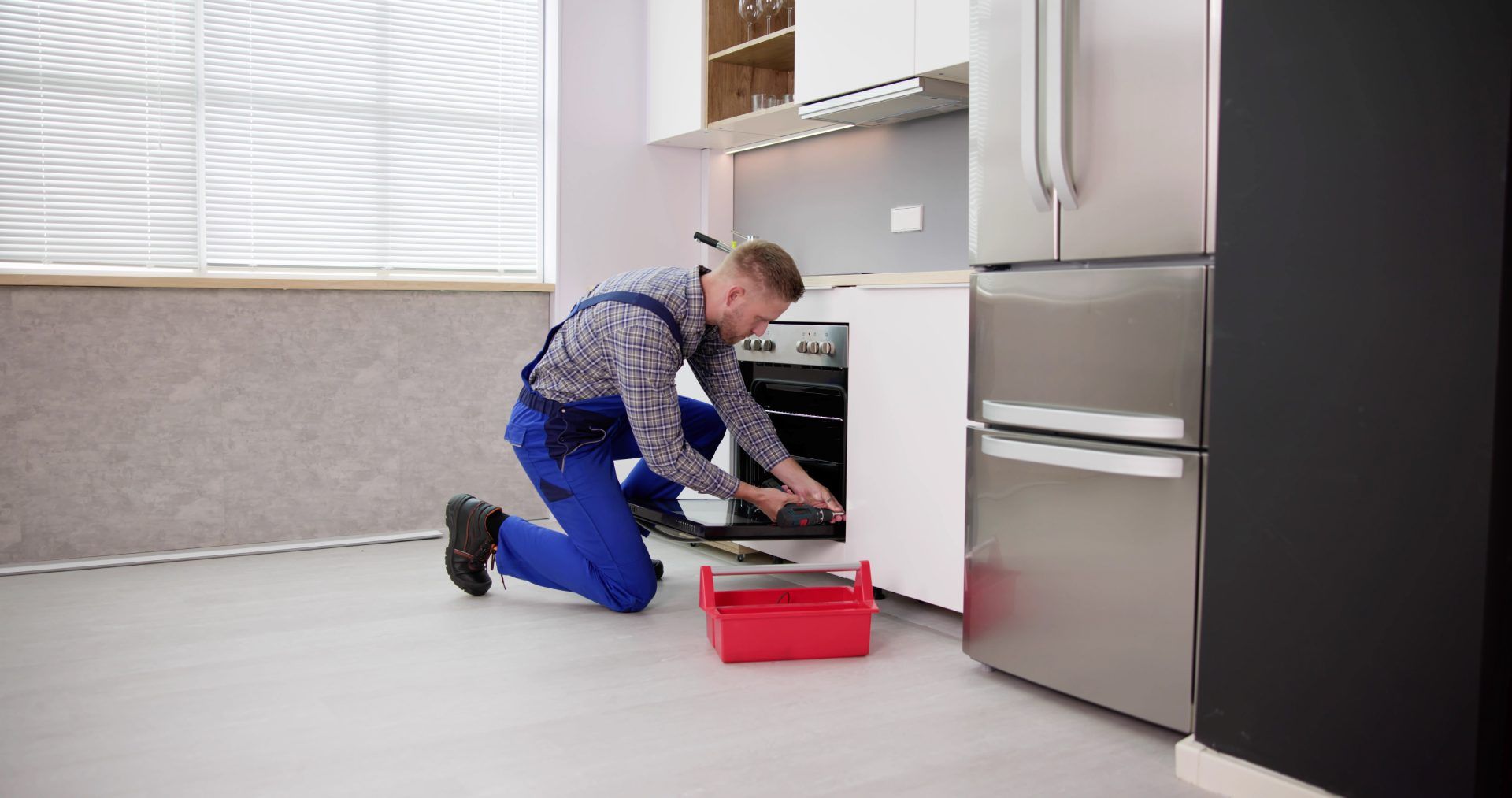 A repairman in blue coveralls kneels to work on an oven in a kitchen. A toolbox sits nearby.