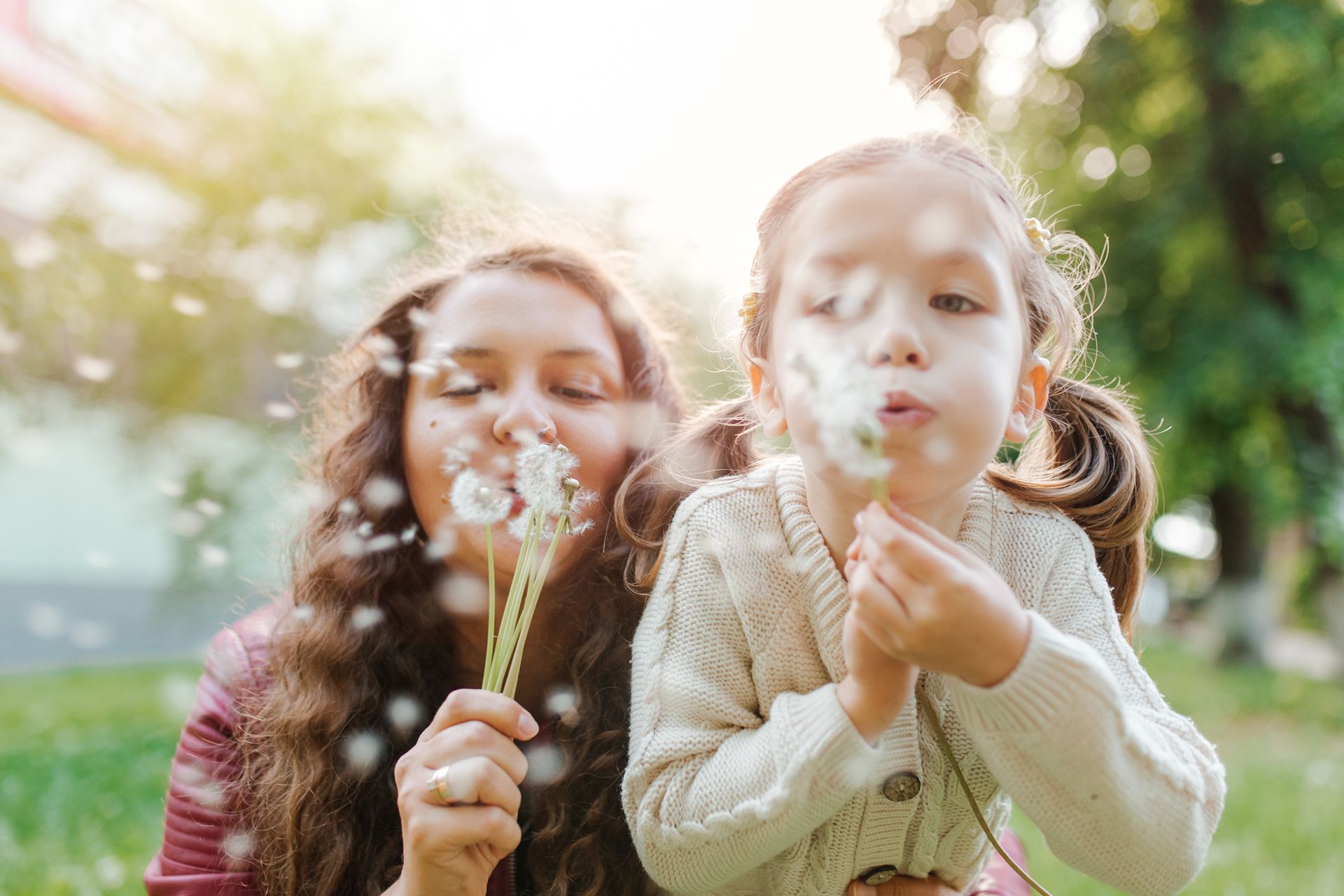 Woman and child blowing dandelion seeds outdoors on a sunny day.