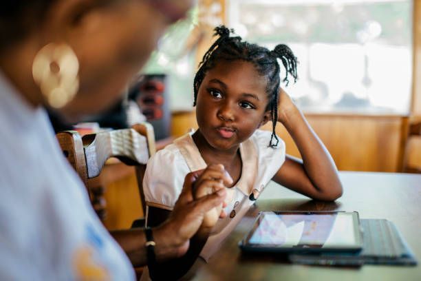 A woman is talking to a little girl who is sitting at a table with a tablet.