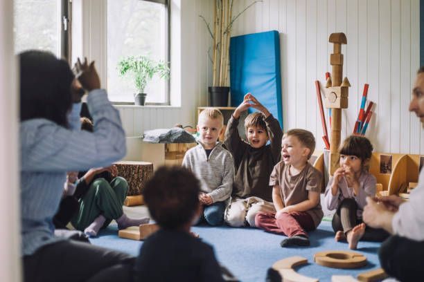 Children seated in a circle, watching adults gesturing in a light-filled classroom.