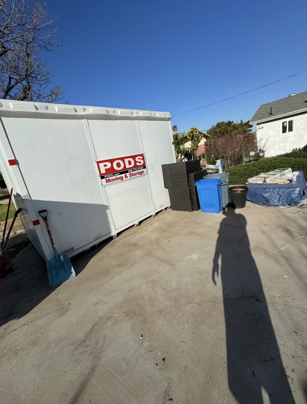 A white PODS container on a concrete driveway, near debris and a house, under a blue sky.