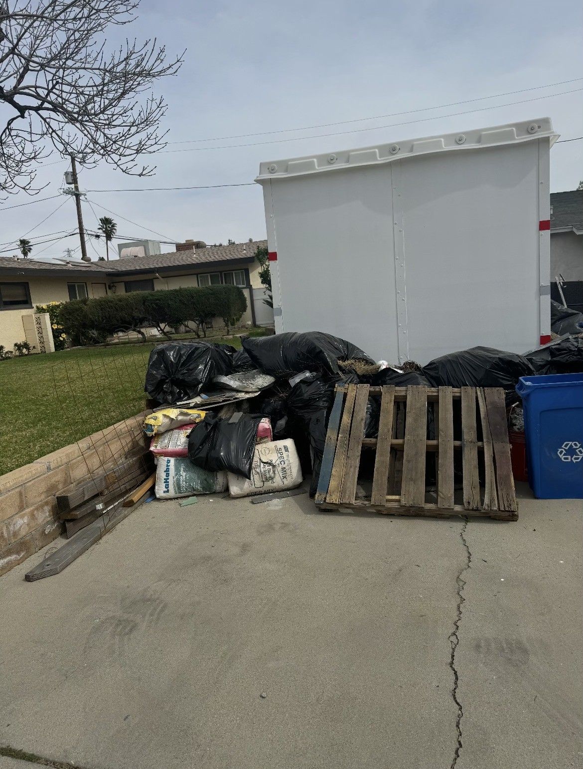 A pile of black trash bags, cardboard, and a wooden pallet are on a driveway.