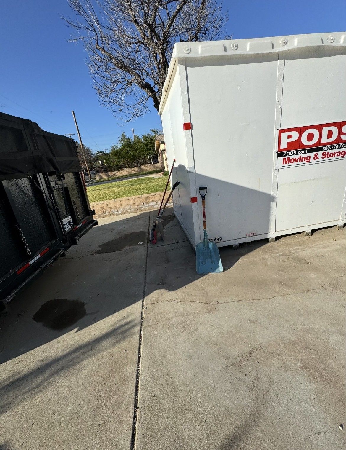 White PODS container next to a black trailer on a concrete surface, shovel leaning against PODS.