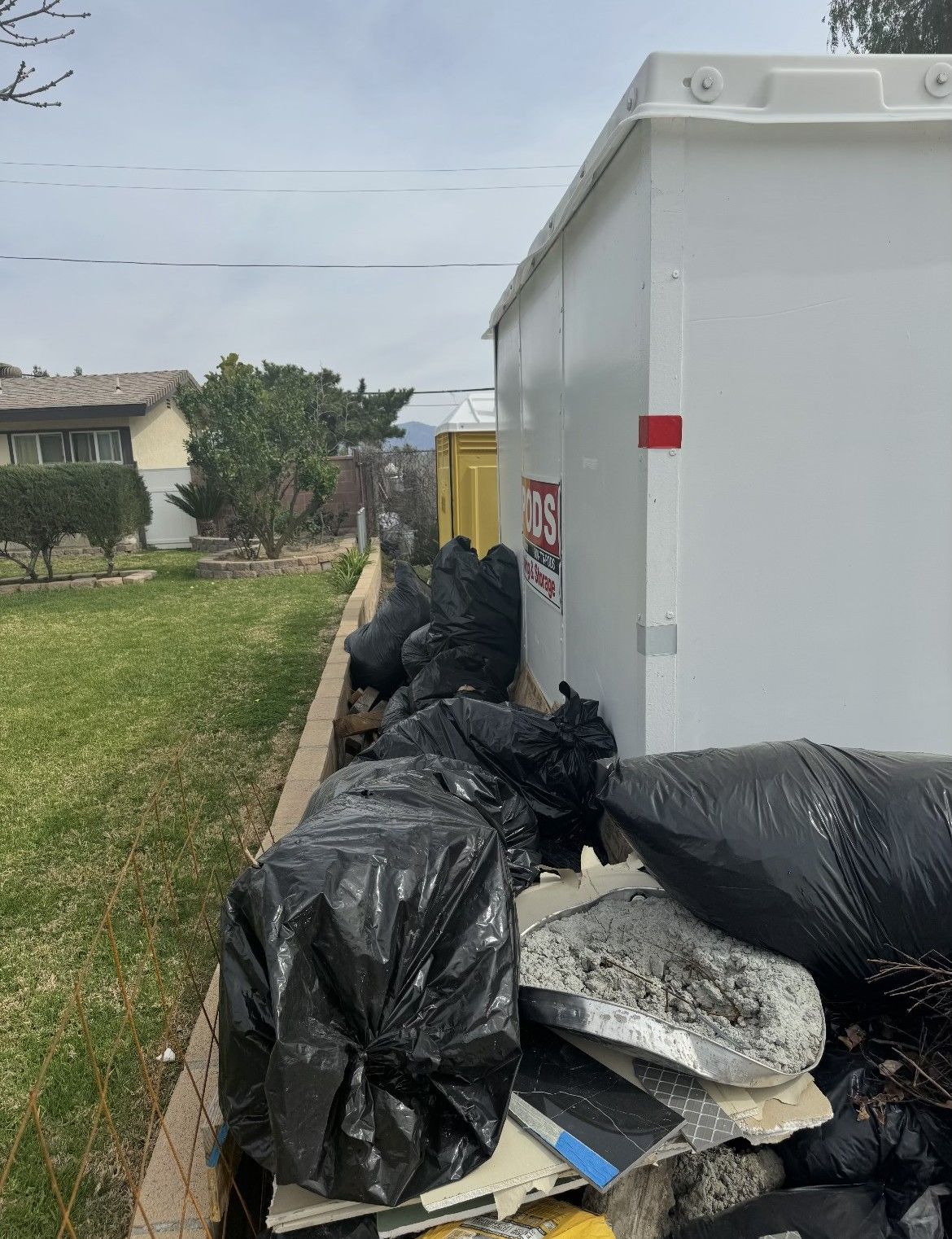 Black trash bags piled near a white dumpster in a grassy yard, near a residential area.