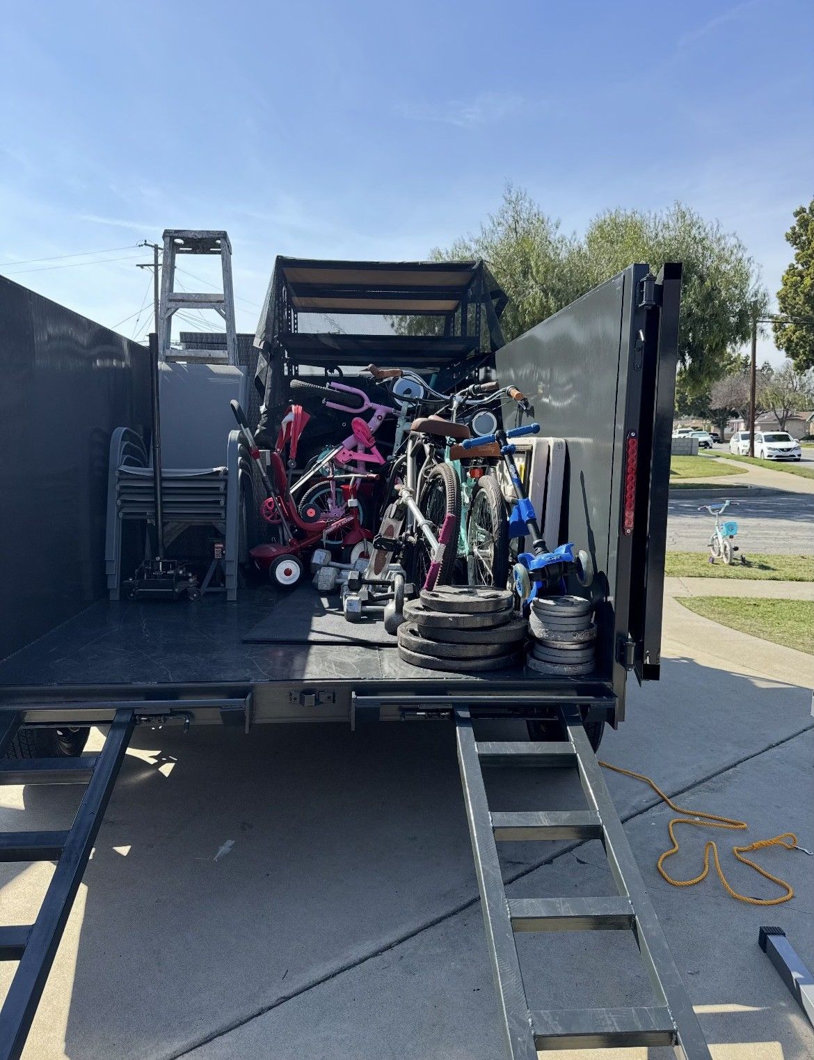 A truck bed loaded with various bikes, weights, and a ladder, parked on a driveway.