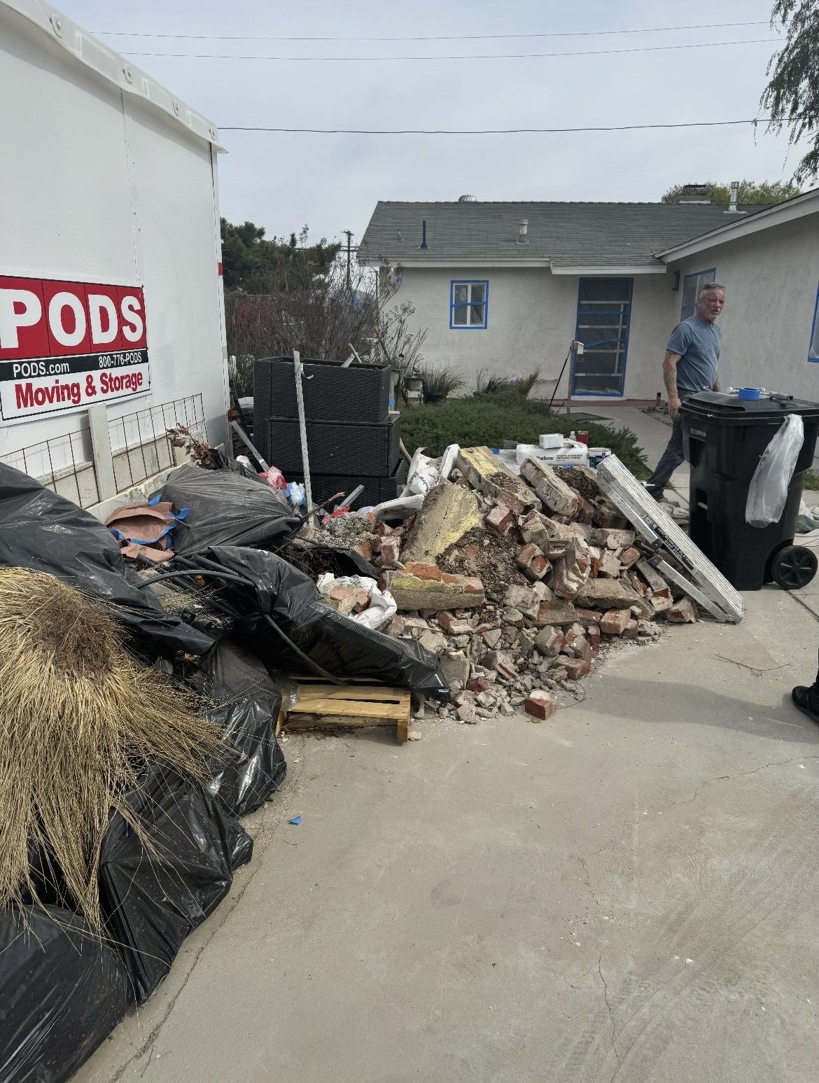 Debris pile of brick and trash next to a PODS container in a driveway, person walks near a house.