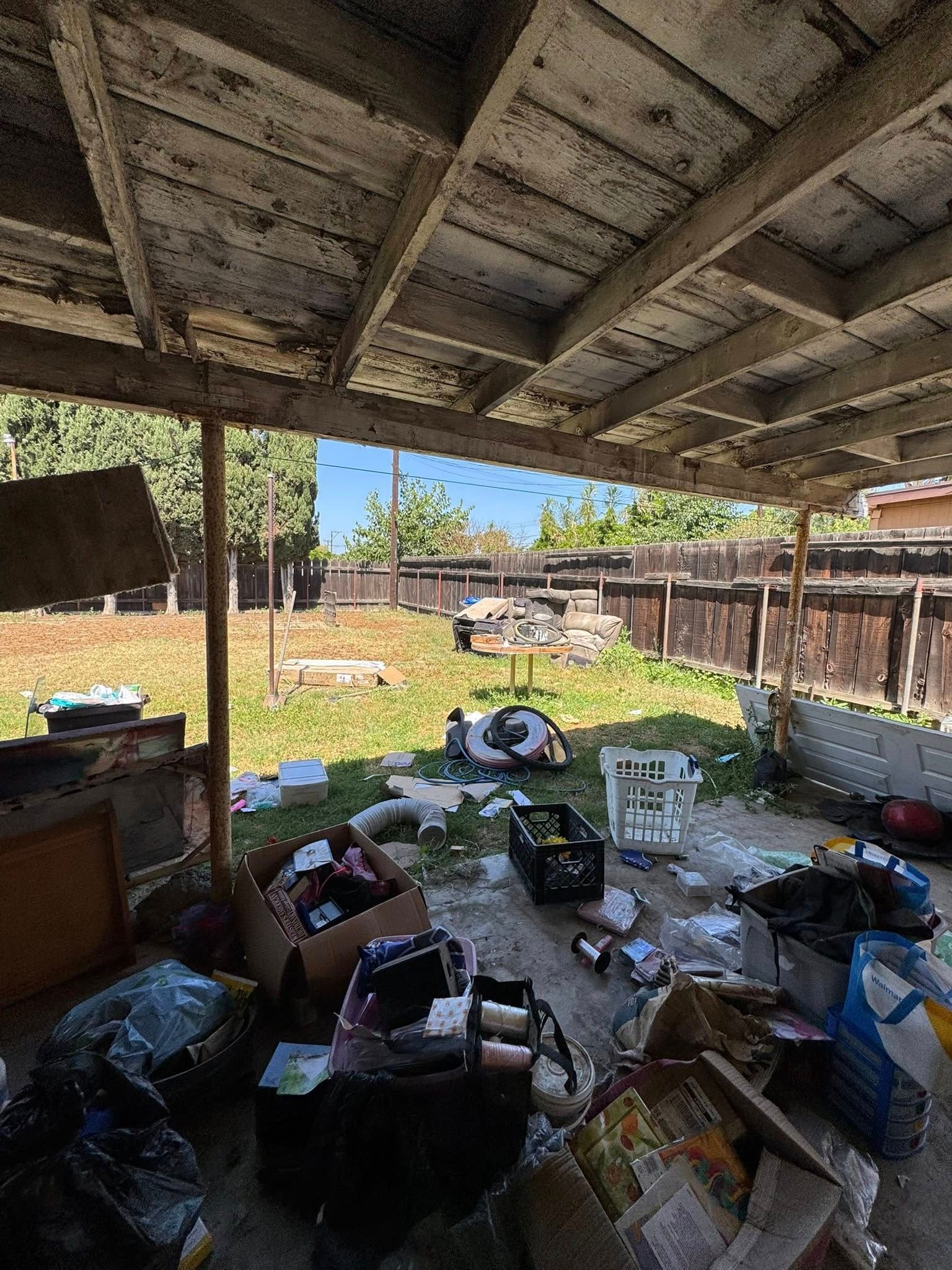 A cluttered backyard under a deteriorating wooden patio cover. Trash and debris cover the ground.