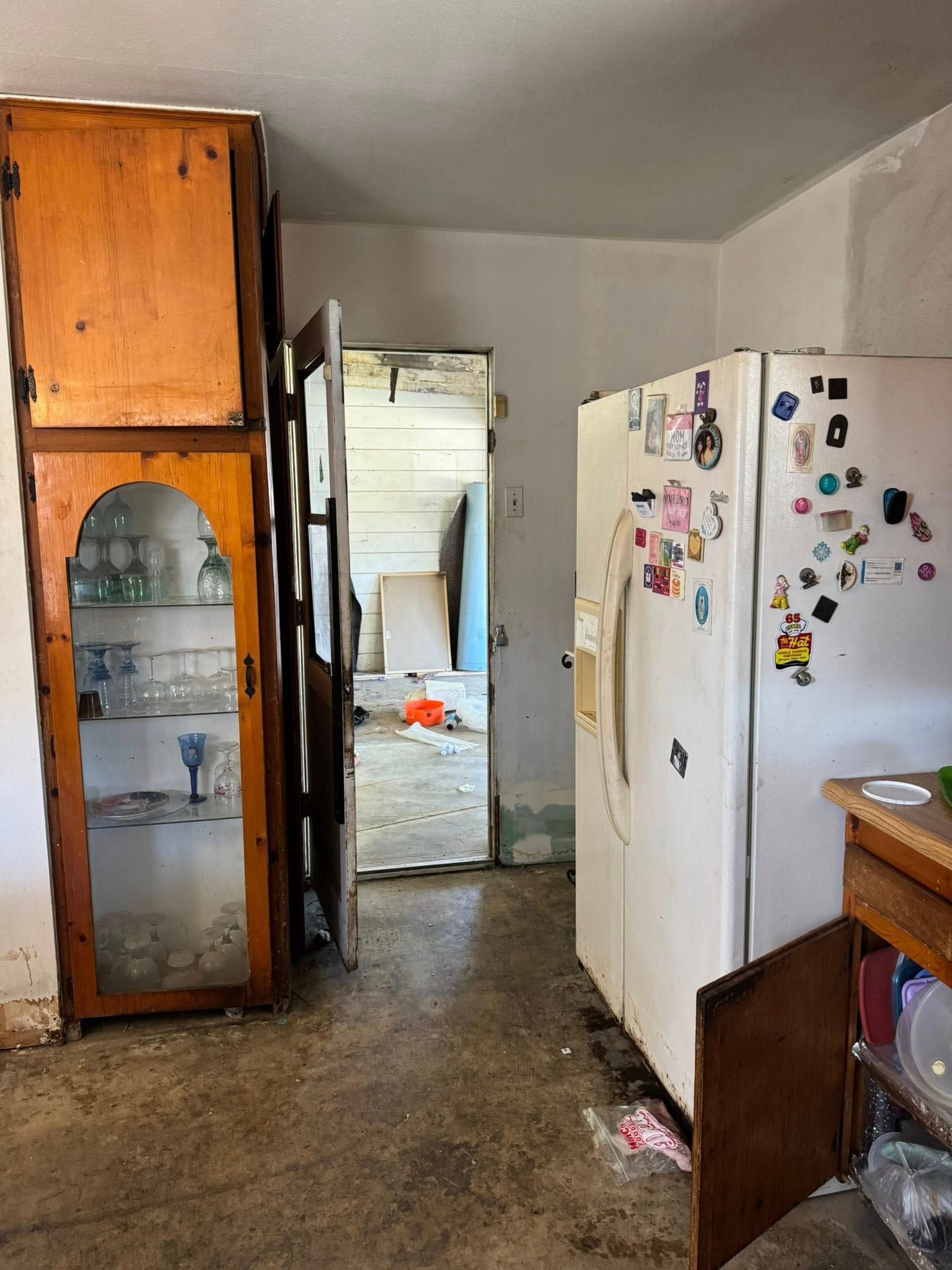 A cluttered kitchen with stained floors, cabinets, refrigerator, and doorway leading to a messy room.