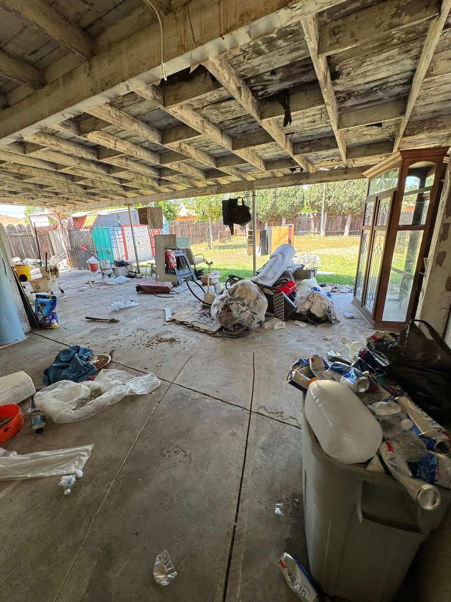 A cluttered outdoor patio. Debris and trash litter the concrete floor under a weathered ceiling. Backyard visible.
