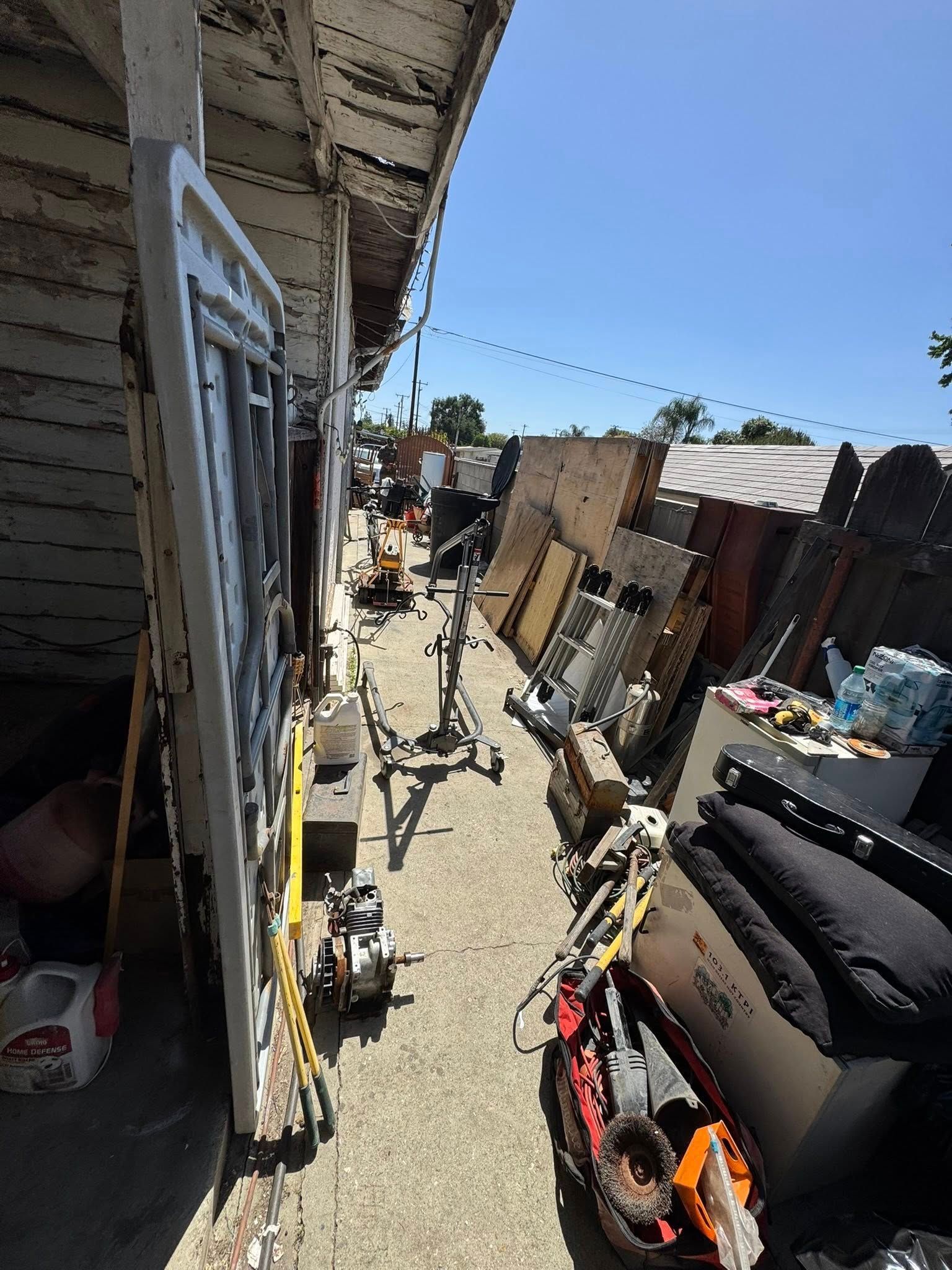 Narrow outdoor alley filled with stored items: doors, lumber, equipment. Bright sunlight.