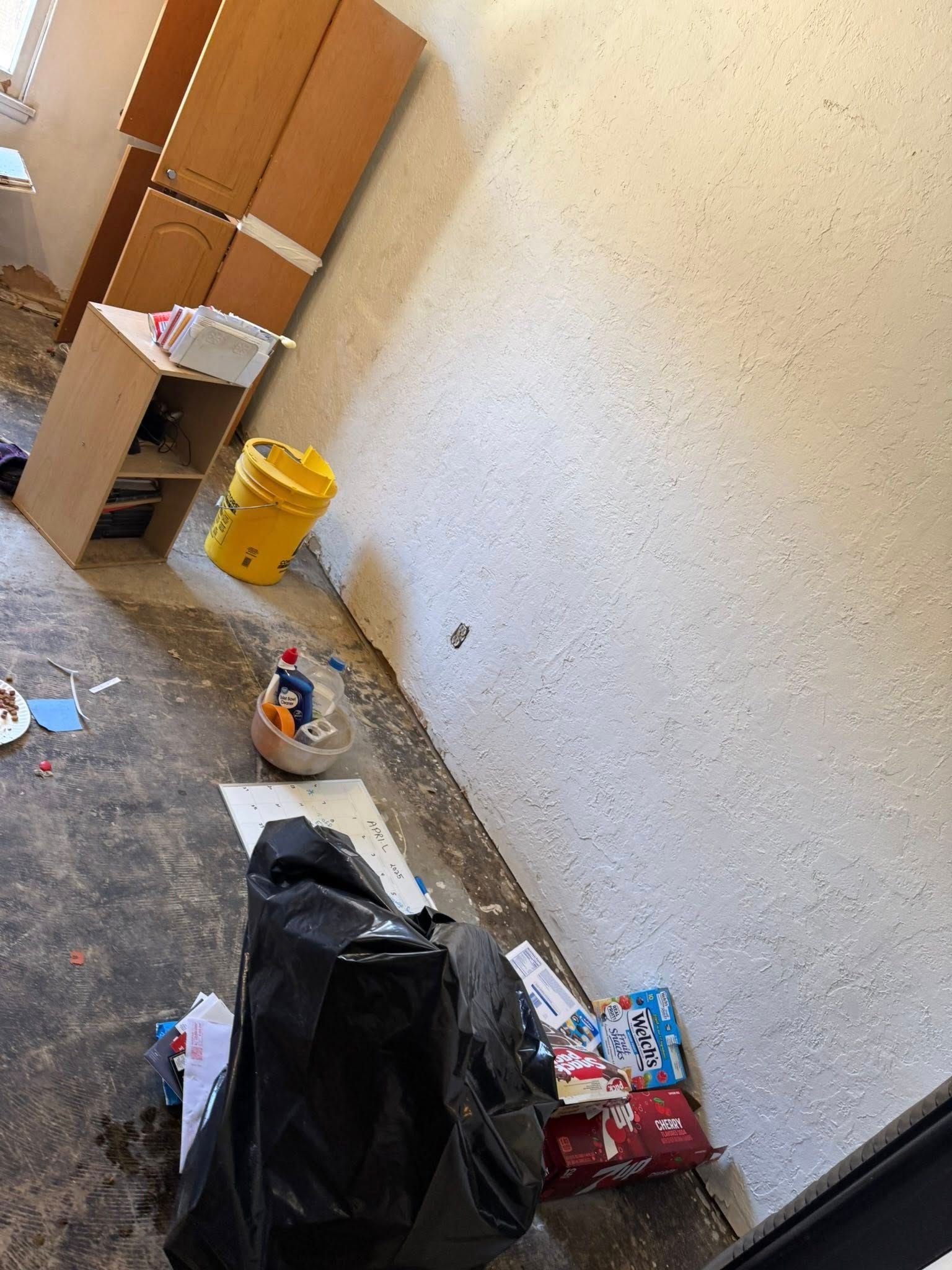 Room with cabinets, debris, and a yellow bucket against a textured wall.