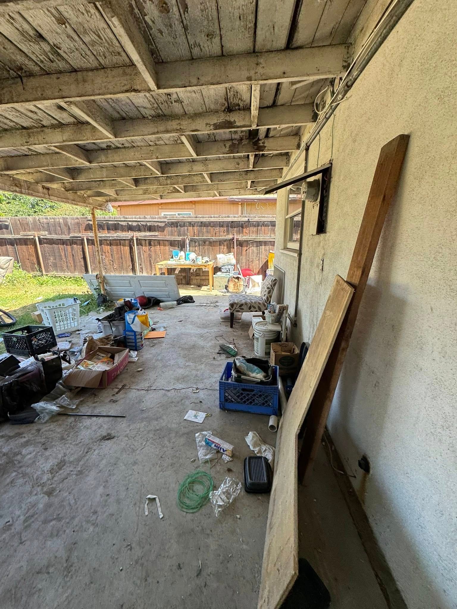 Overgrown cluttered outdoor area with peeling ceiling, debris on ground, and fence in the background.