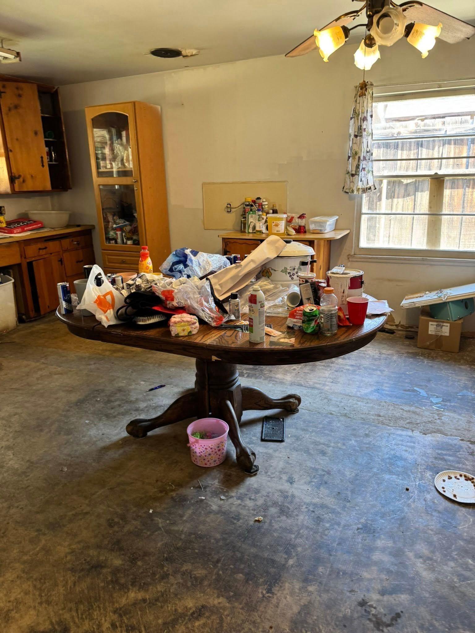 Messy kitchen: Round table piled with items, cabinets, glass-front cabinet, window with curtains. Concrete floor.