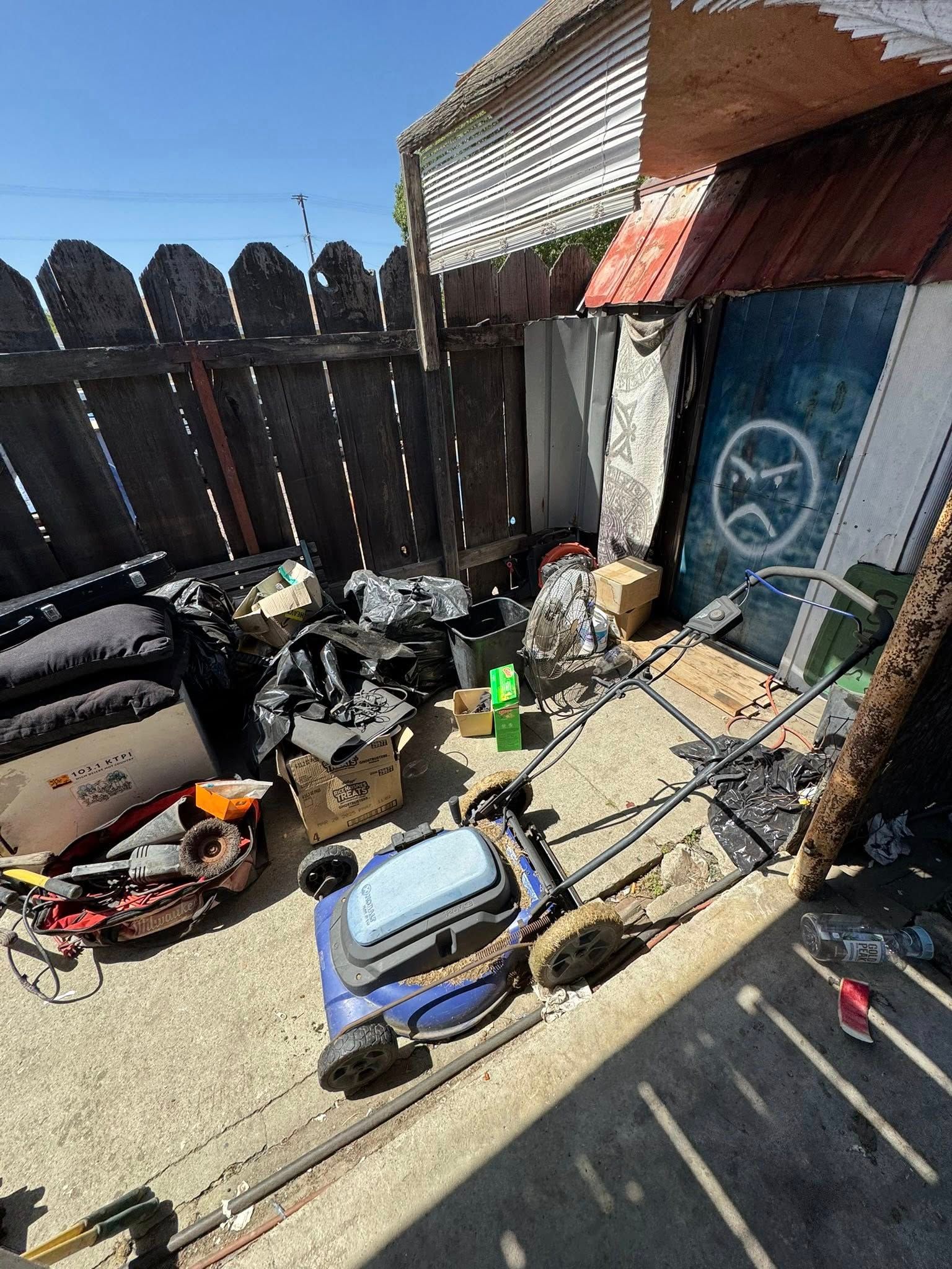 Backyard with a lawnmower, trash, and a shed against a wooden fence under a blue sky.