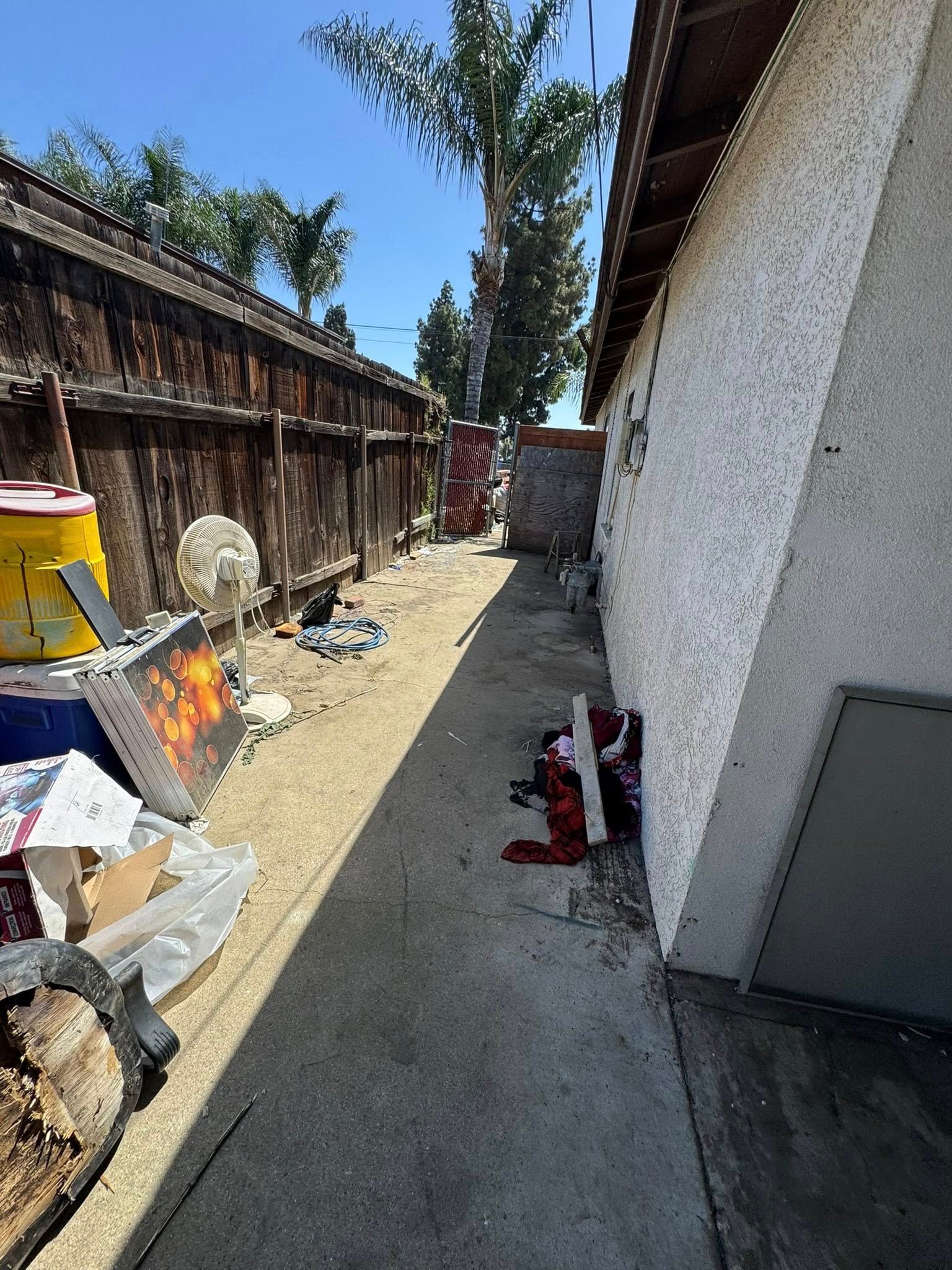 Narrow, debris-filled side yard between a wooden fence and a stucco wall on a sunny day.