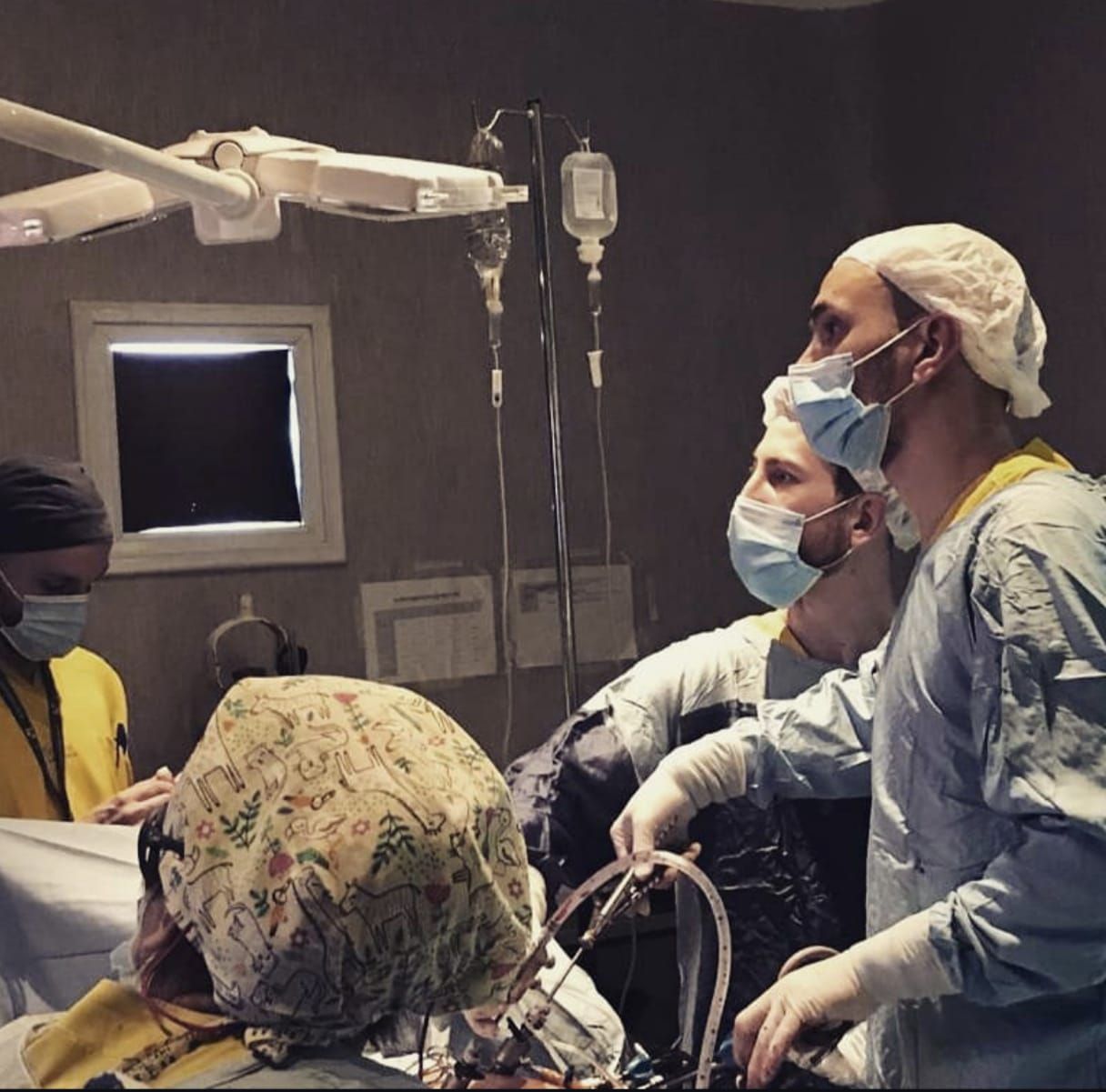 Equipo médico en un quirófano, observando una cirugía. Se ven lámparas, equipo médico y rostros con mascarillas.