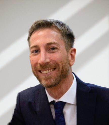Hombre de pelo corto y castaño, con barba, sonriente, vestido con un traje oscuro y una corbata azul.