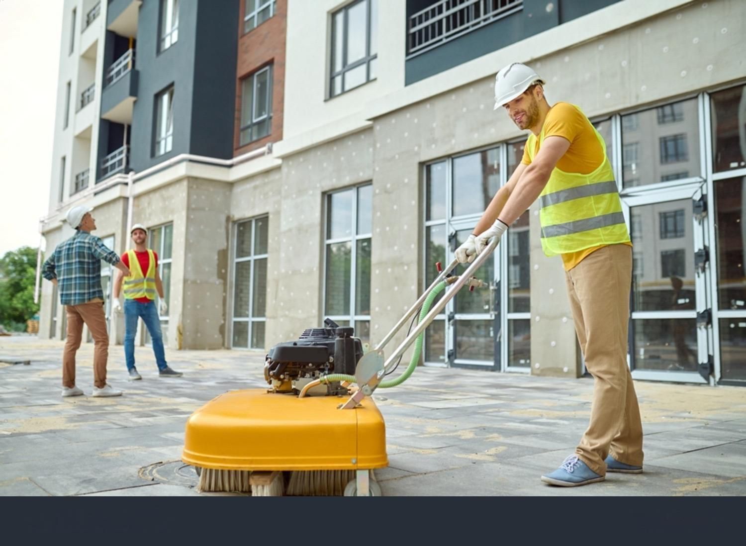 Construction worker using a power broom on a concrete surface outside a building. Two other workers watch.