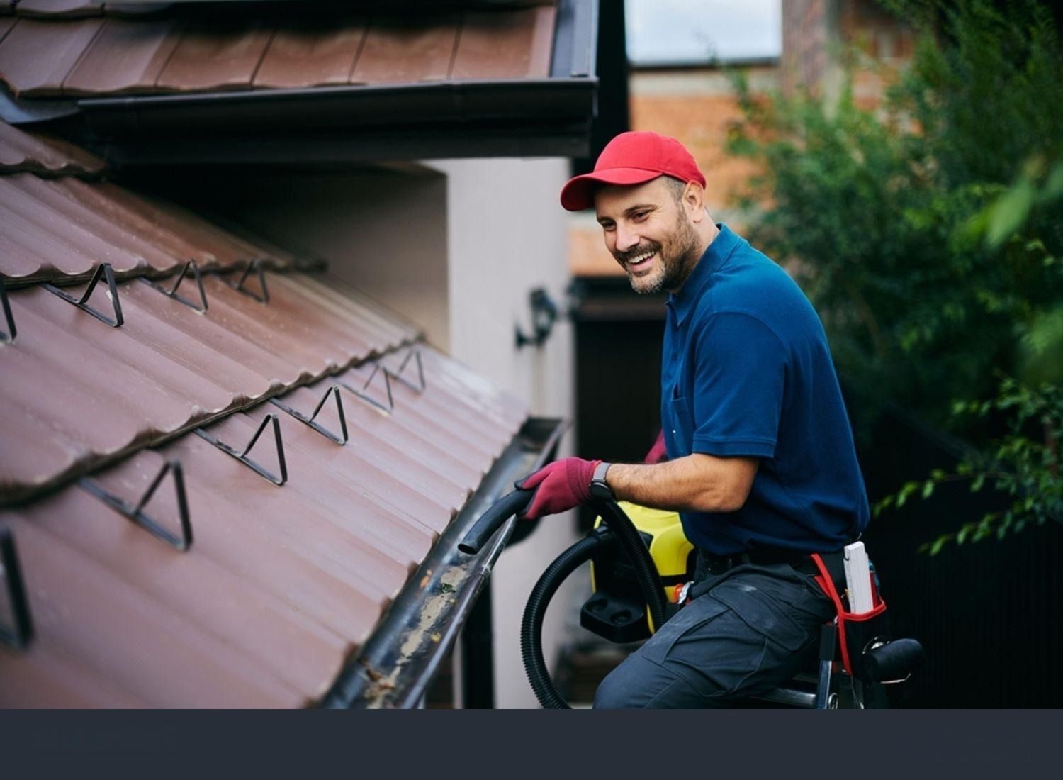 Man cleaning a roof gutter, smiling, wearing a red hat and gloves, holding a cleaning tool.