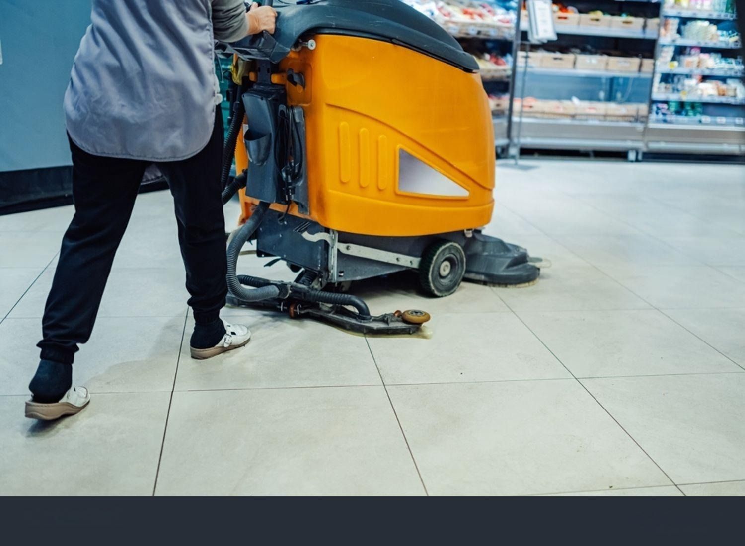 Person using an orange floor cleaning machine in a store aisle with tile flooring.
