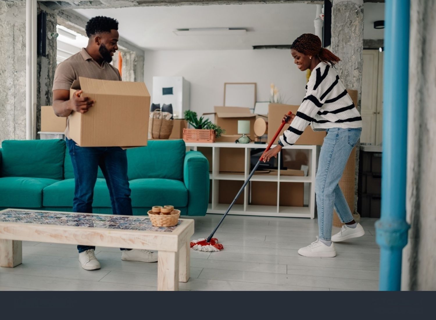 Couple moving into a new home. Man carries a box, woman sweeps floor. Bright living room.