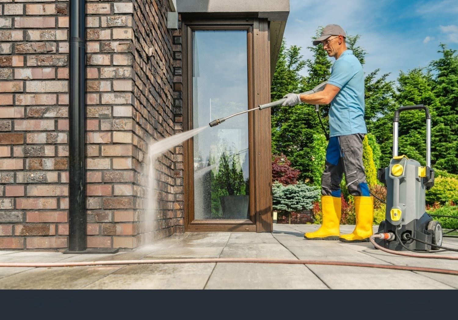 Man power washing the exterior of a brick building. He wears a hat, boots and gloves.
