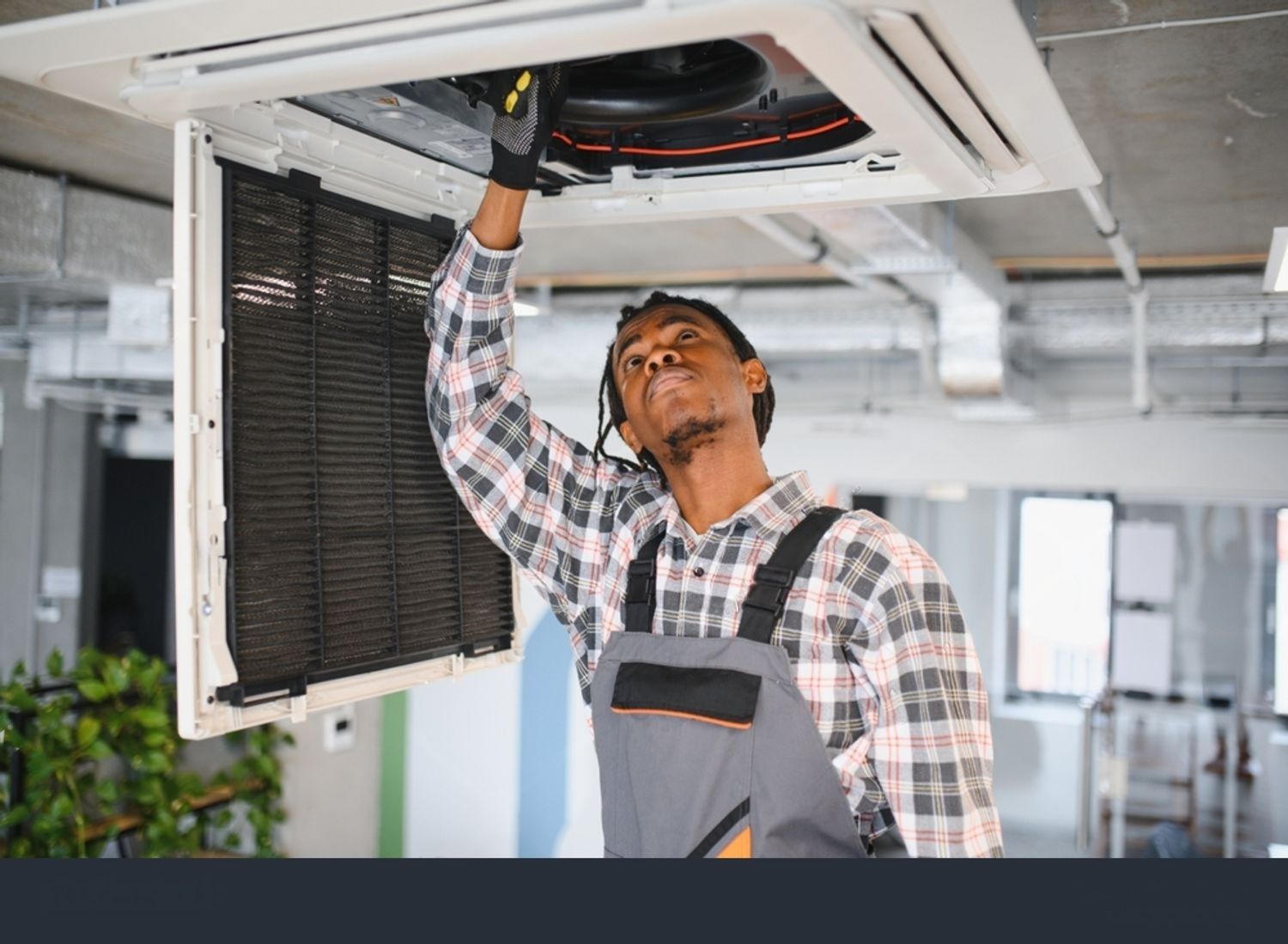 A person in overalls repairs an air conditioning unit in an office.