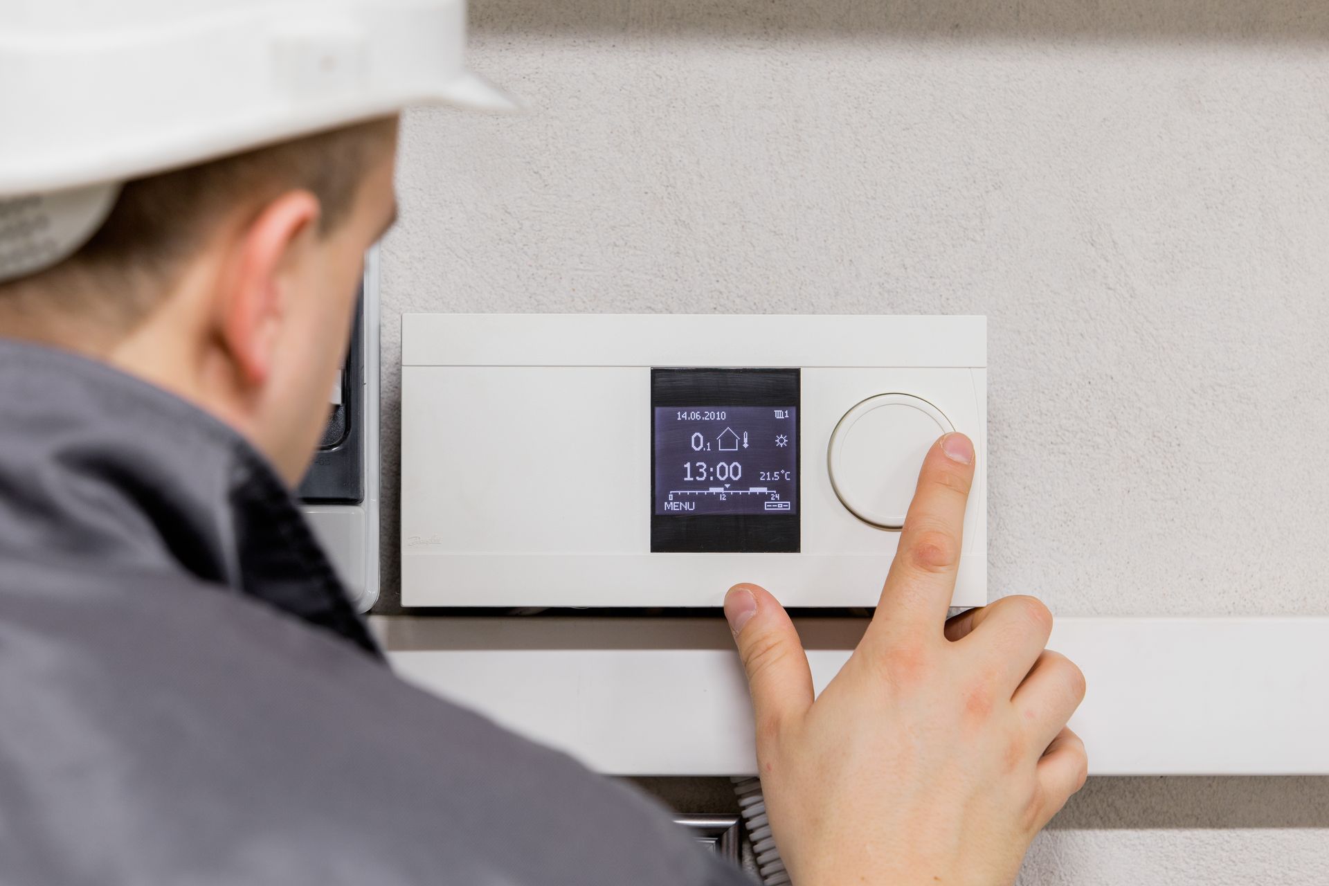 A person in a hard hat adjusts a thermostat mounted on a white wall.