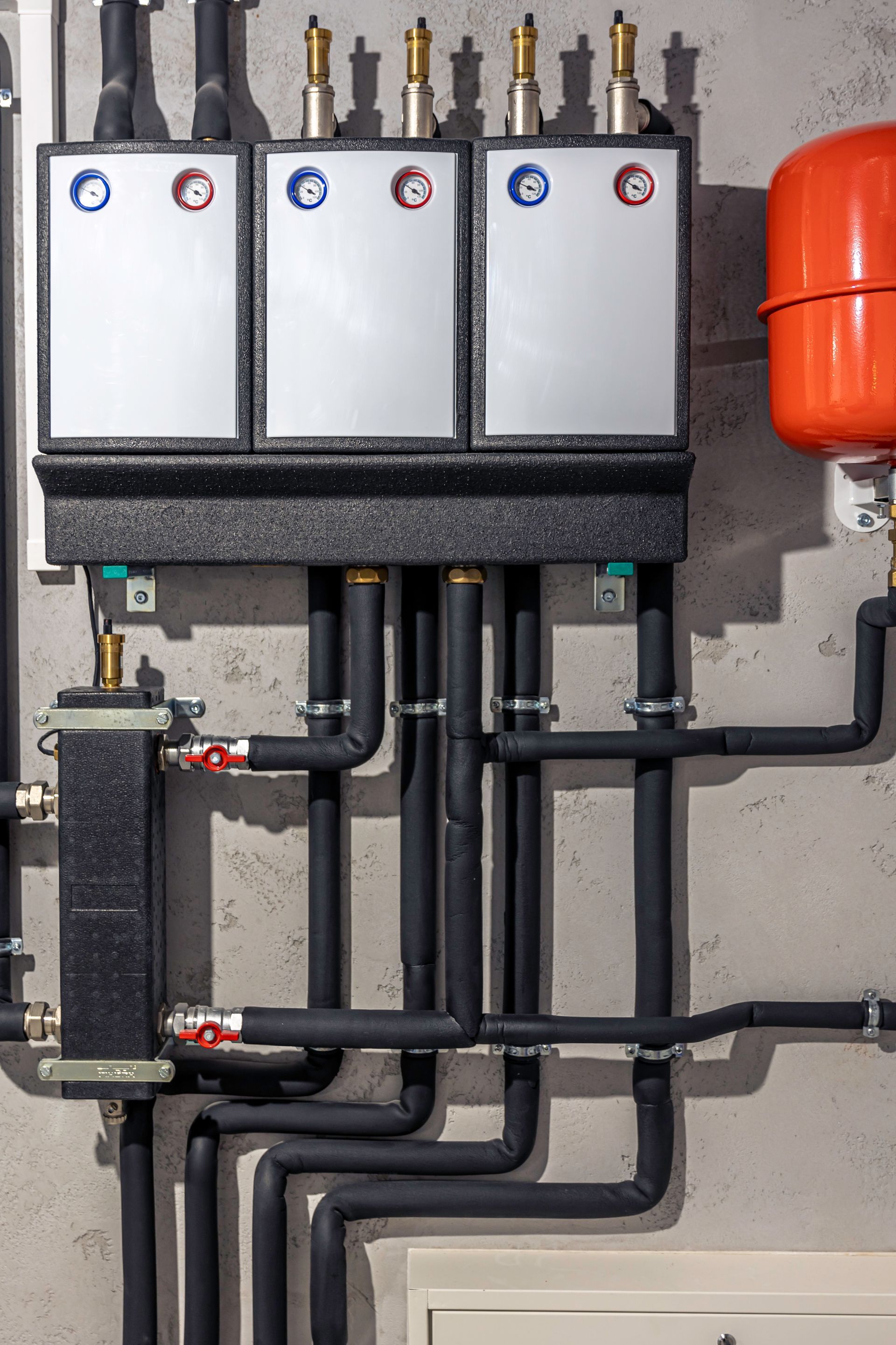 Plumbing system with black pipes, silver panels, and an orange tank against a gray wall.