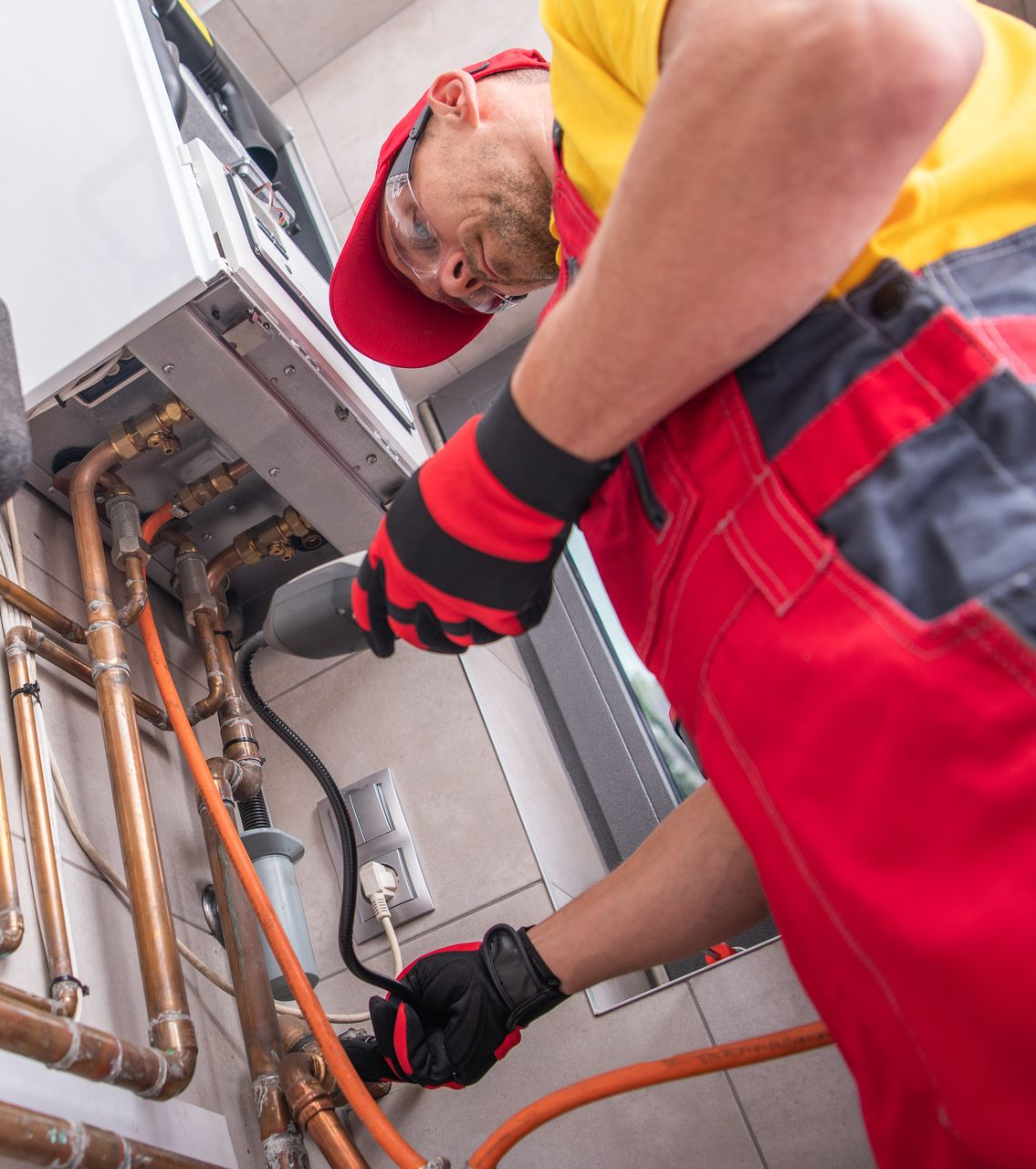 Plumber in red overalls and hat working on copper pipes connected to a heating unit.