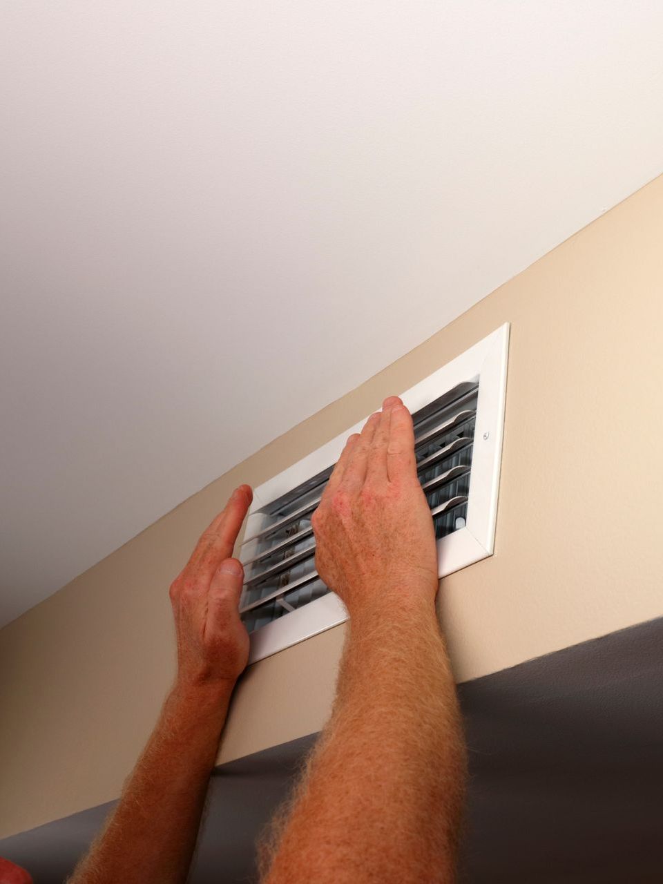 Hands removing a white air vent cover from a tan wall, against a white ceiling.