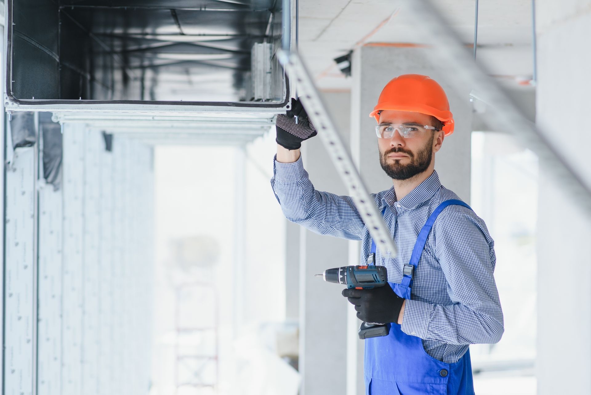 Construction worker in orange hard hat, safety glasses, and blue overalls, using a drill on ductwork.