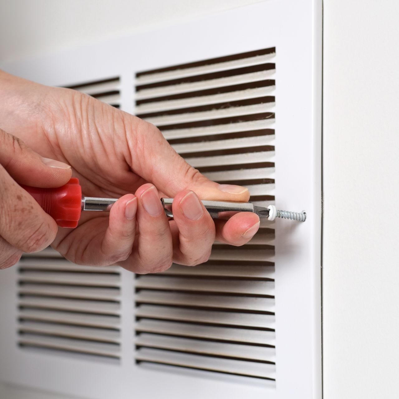 Person using a screwdriver to unscrew a panel with vent slats. The panel is white.