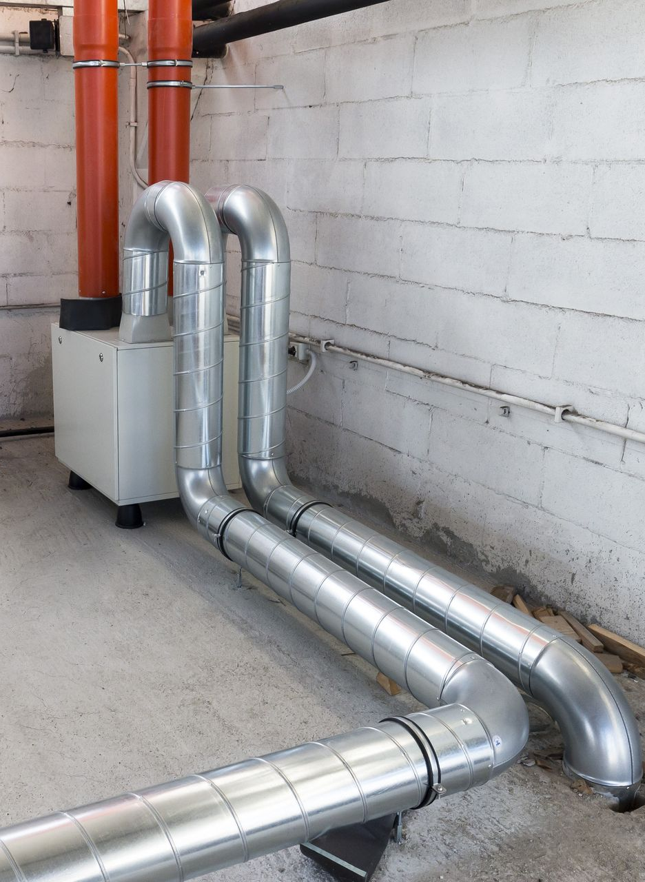Metal ductwork connected to a rectangular appliance in a concrete room with red and black pipes.
