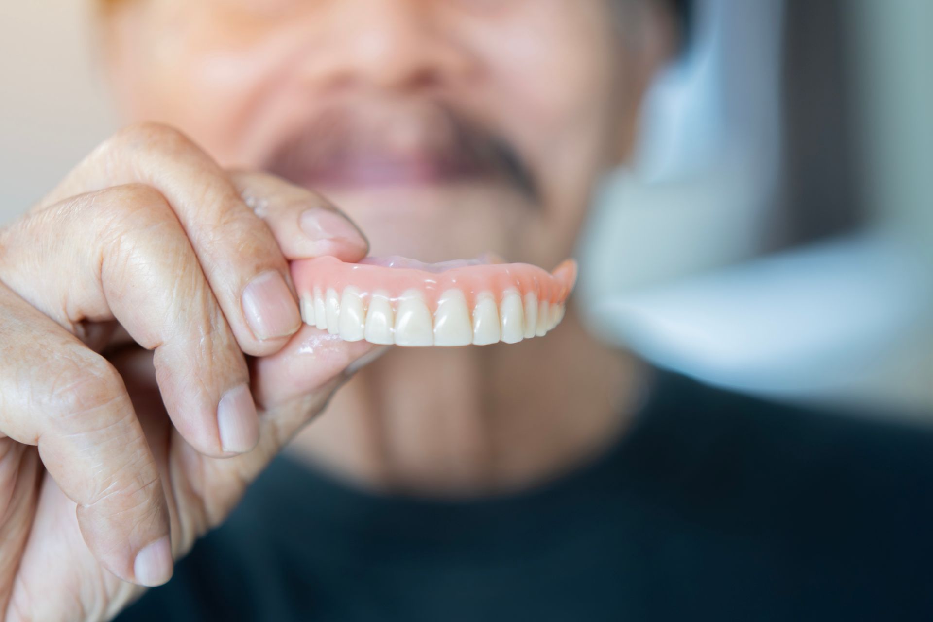 Waist-up portrait of a senior man with a mustache wearing a black t-shirt holding denture implants.
