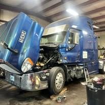 Blue semi-truck with the hood open in a repair shop, showing engine components.