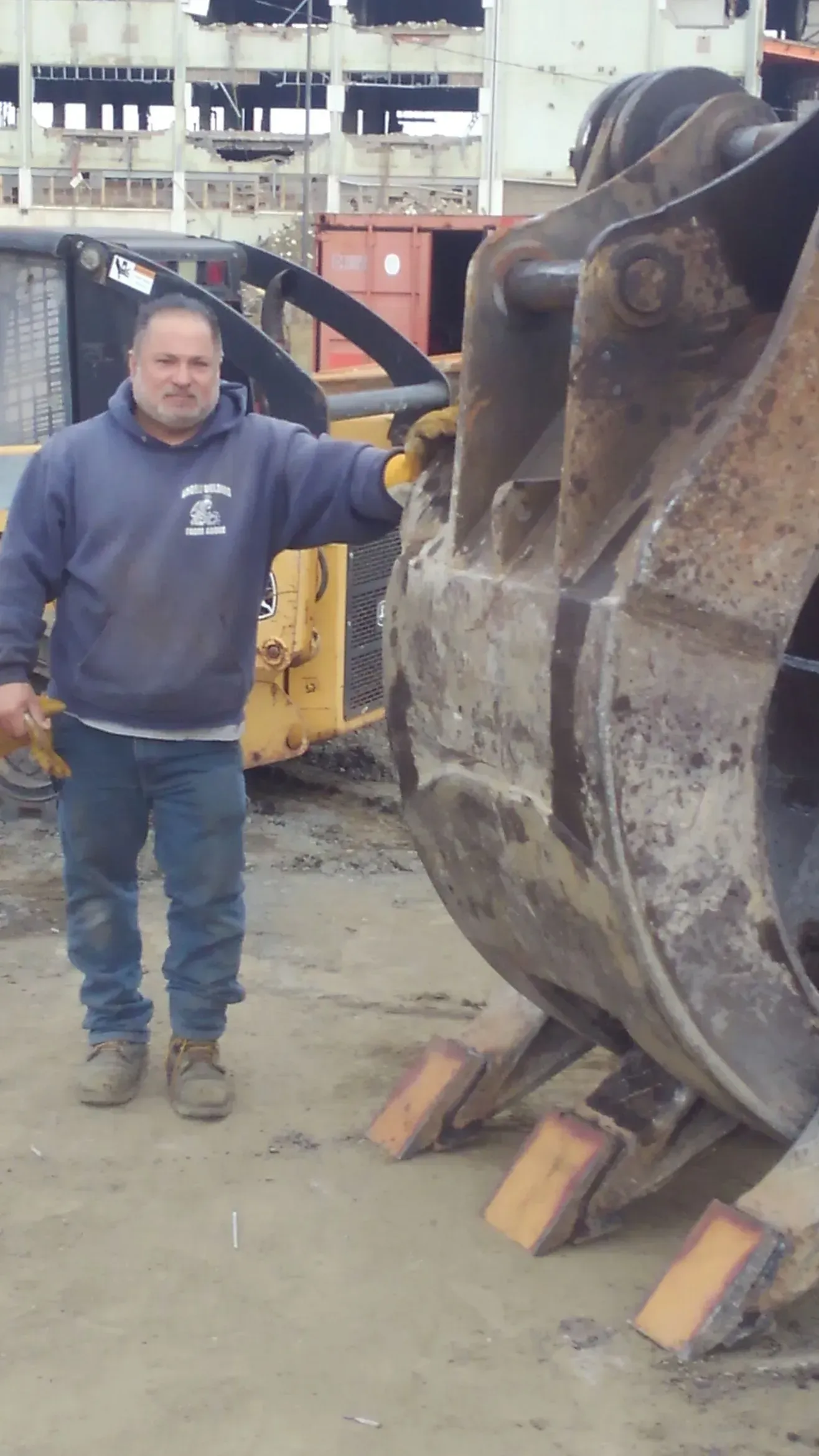 A man in a blue sweatshirt stands next to a large excavator bucket