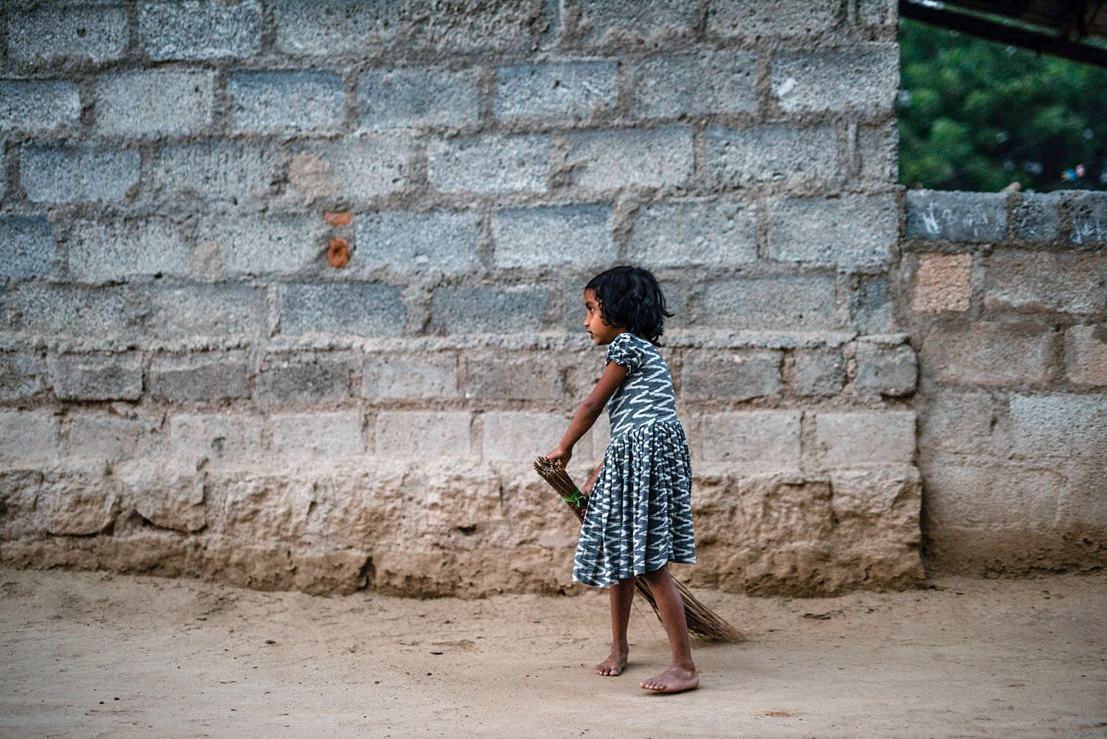 A little girl is sweeping the ground in front of a brick wall.
