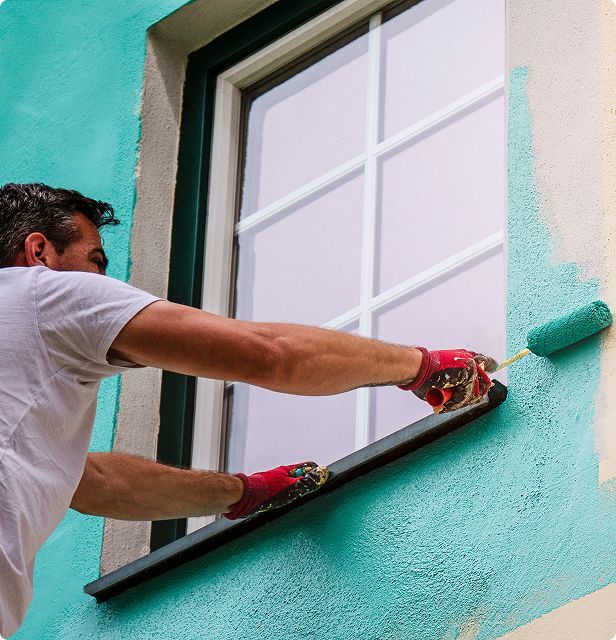A person paints a teal wall with a roller next to a window. Wearing red gloves, the painter is outside on a building.