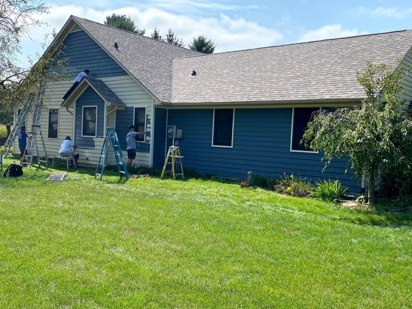 Blue house with steep roof and workers on a front porch in a sunny grassy yard