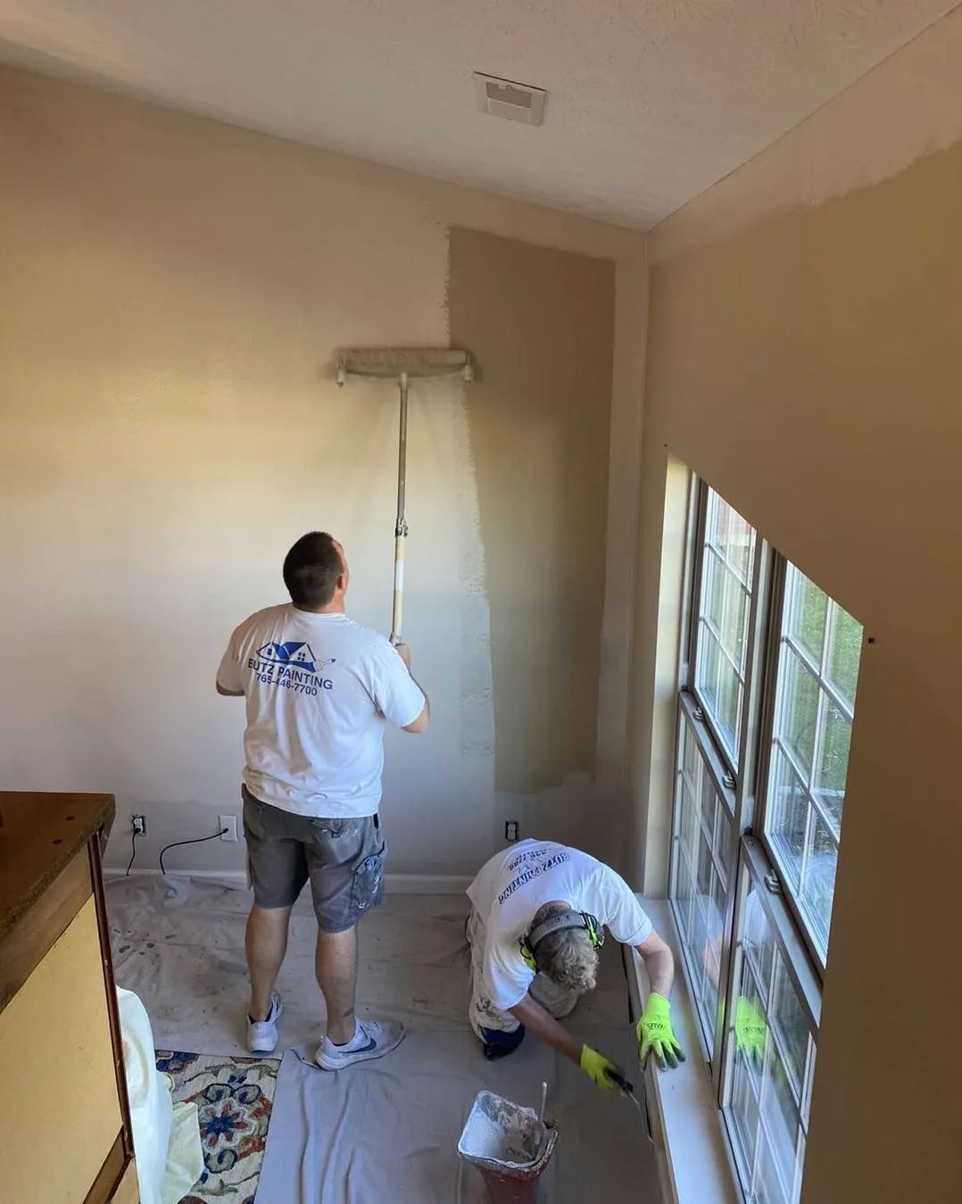 Two workers painting a beige room, one on a ladder near a window and one crouching by supplies.
