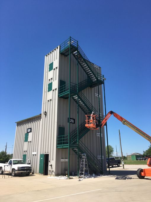 Worker in orange lift beside a tall training tower with exterior stairs under a clear blue sky