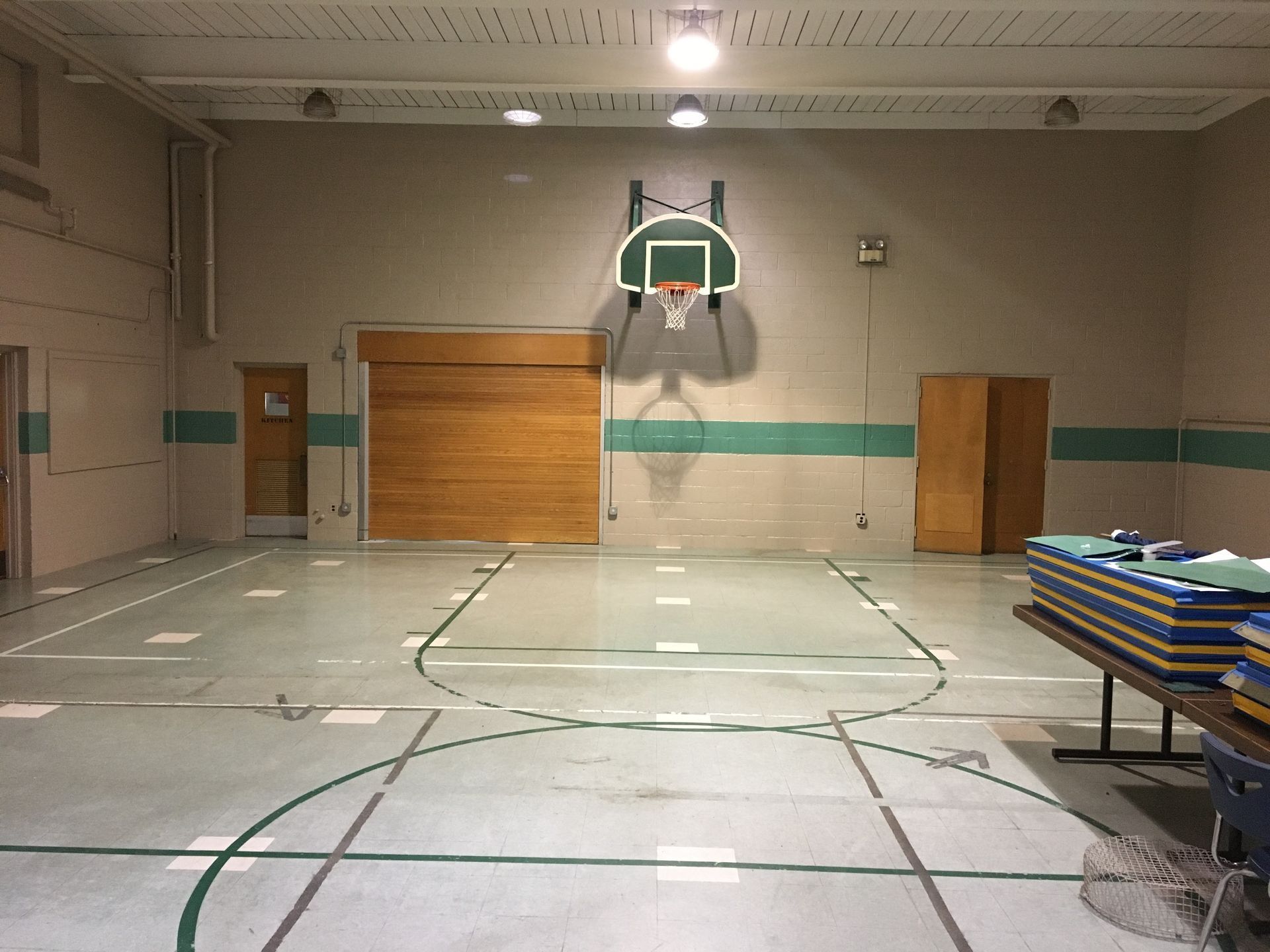 Empty gymnasium with basketball hoop and court lines on a light gray floor, wooden doors at the back.