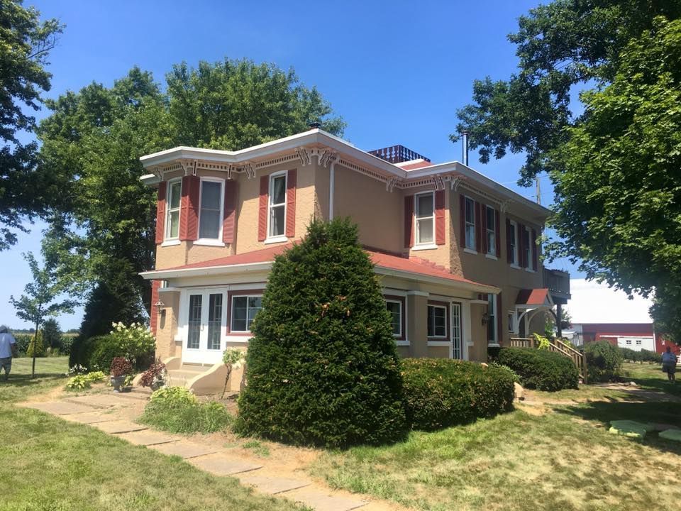 Two-story peach house with white trim, red shutters, and a large evergreen tree in a sunny yard