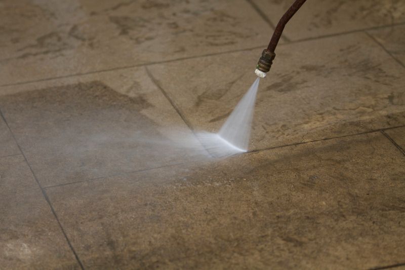 A power washer spraying a stream of water onto dirty tan floor tiles to reveal a cleaner surface.