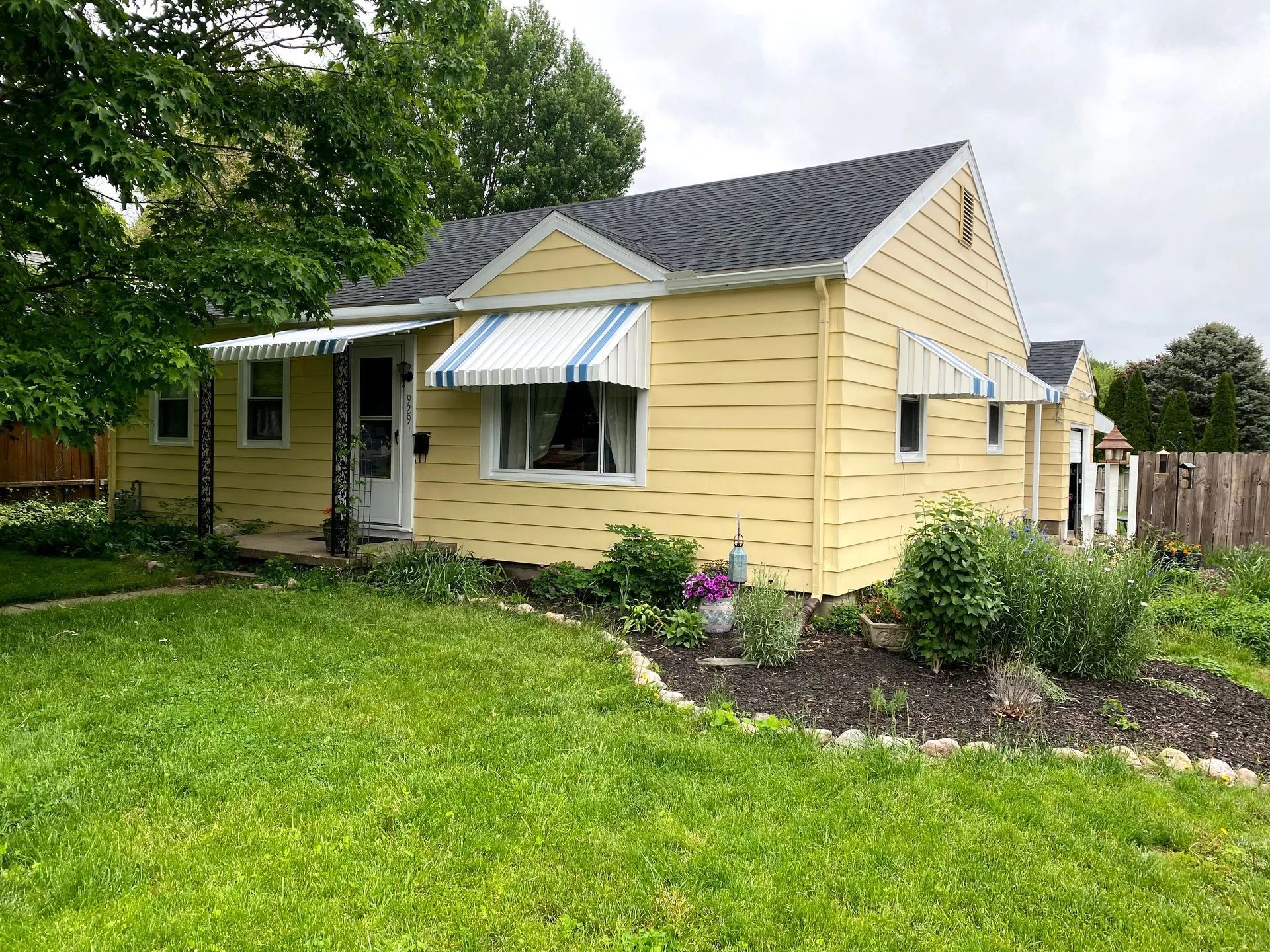 Yellow house with blue and white striped awnings over windows, surrounded by green grass and dark mulch.