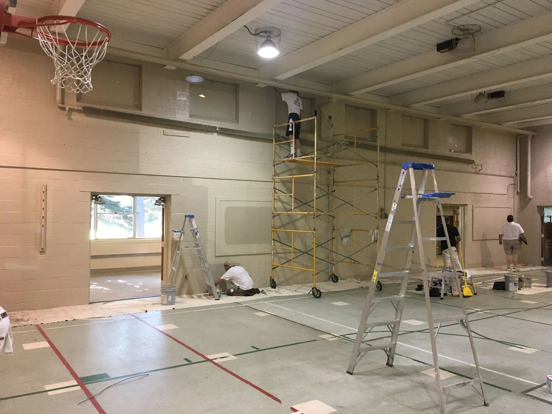 Gymnasium interior during renovation: Workers on scaffolding and ladders, painting walls.
